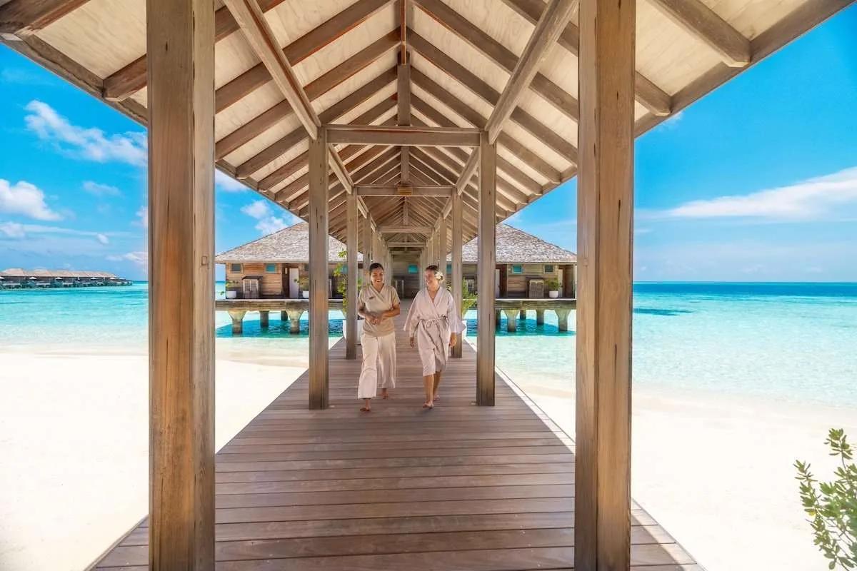 Two guests walking the covered overwater walkway to Sulha Spa's treatment pavilions, one in a robe, beach and lagoon on either side