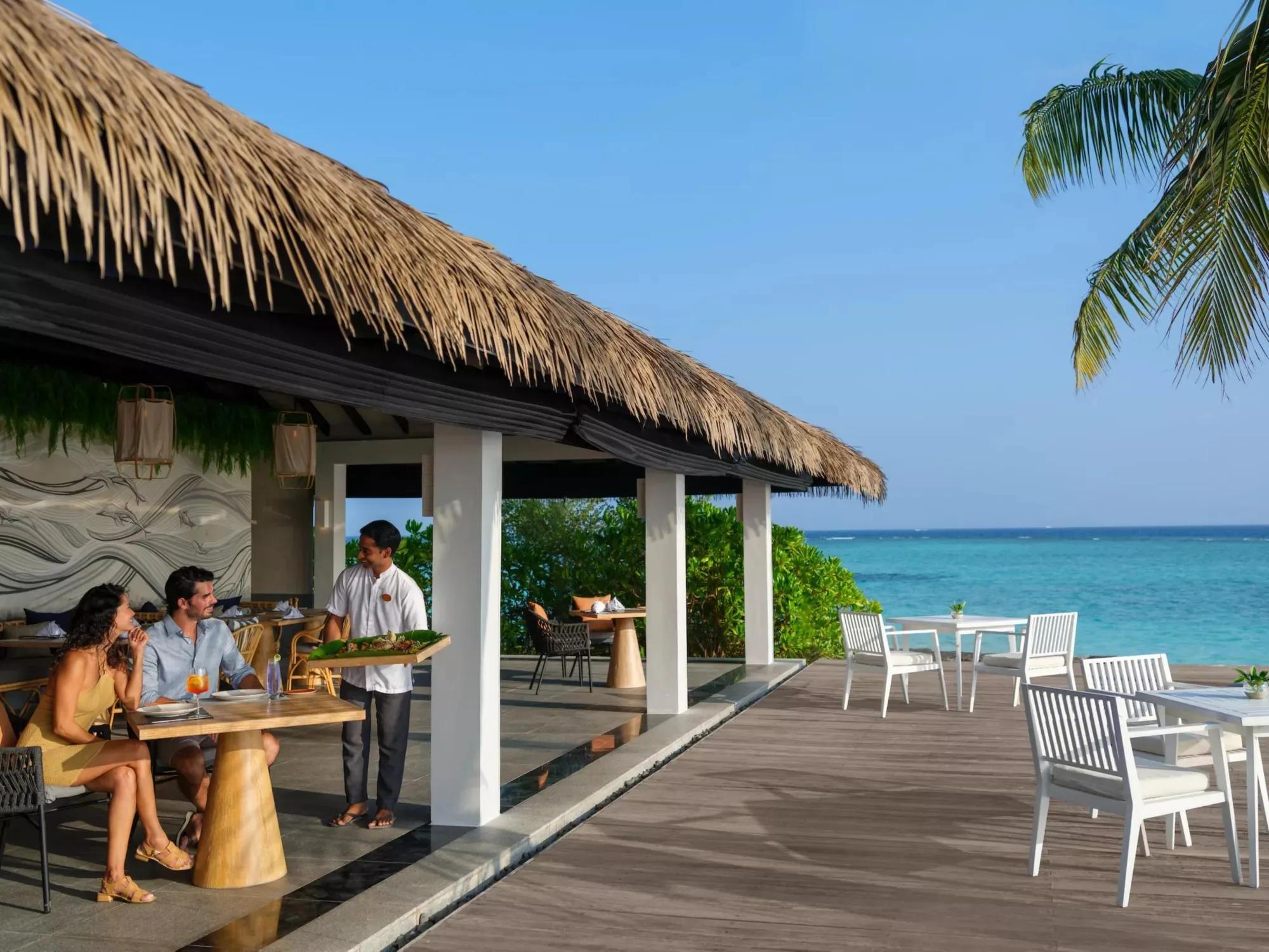 A couple served drinks and food on Palms Bar's beachfront terrace at Noku Maldives