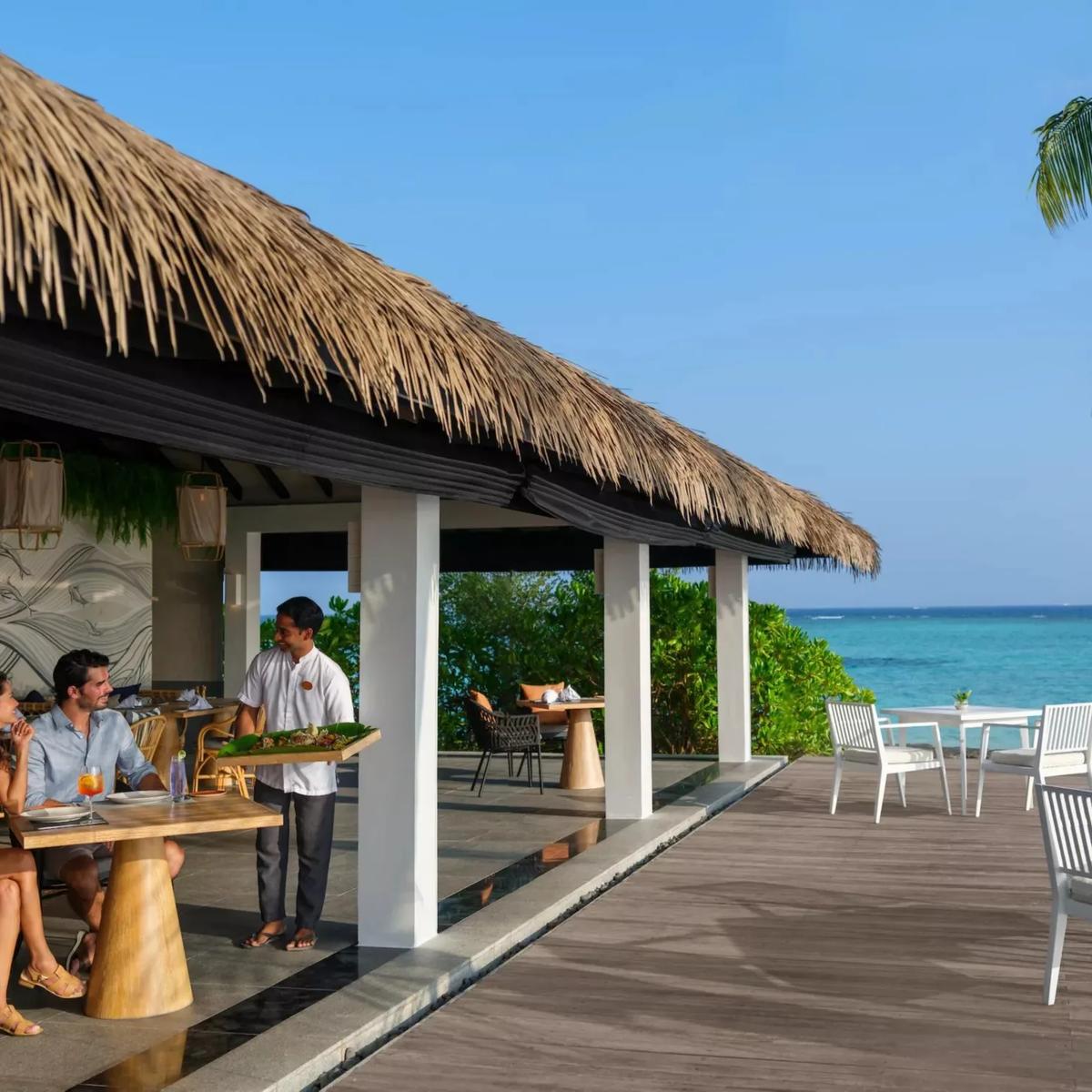 A couple served drinks and food on Palms Bar's beachfront terrace at Noku Maldives