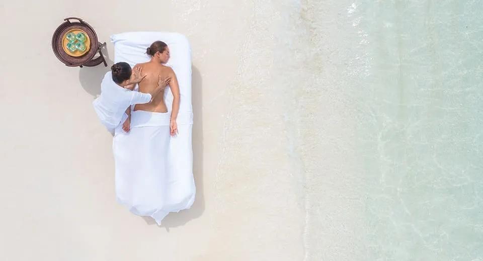 Aerial view of a massage in progress on a beachside daybed, clear shallow water a few metres away