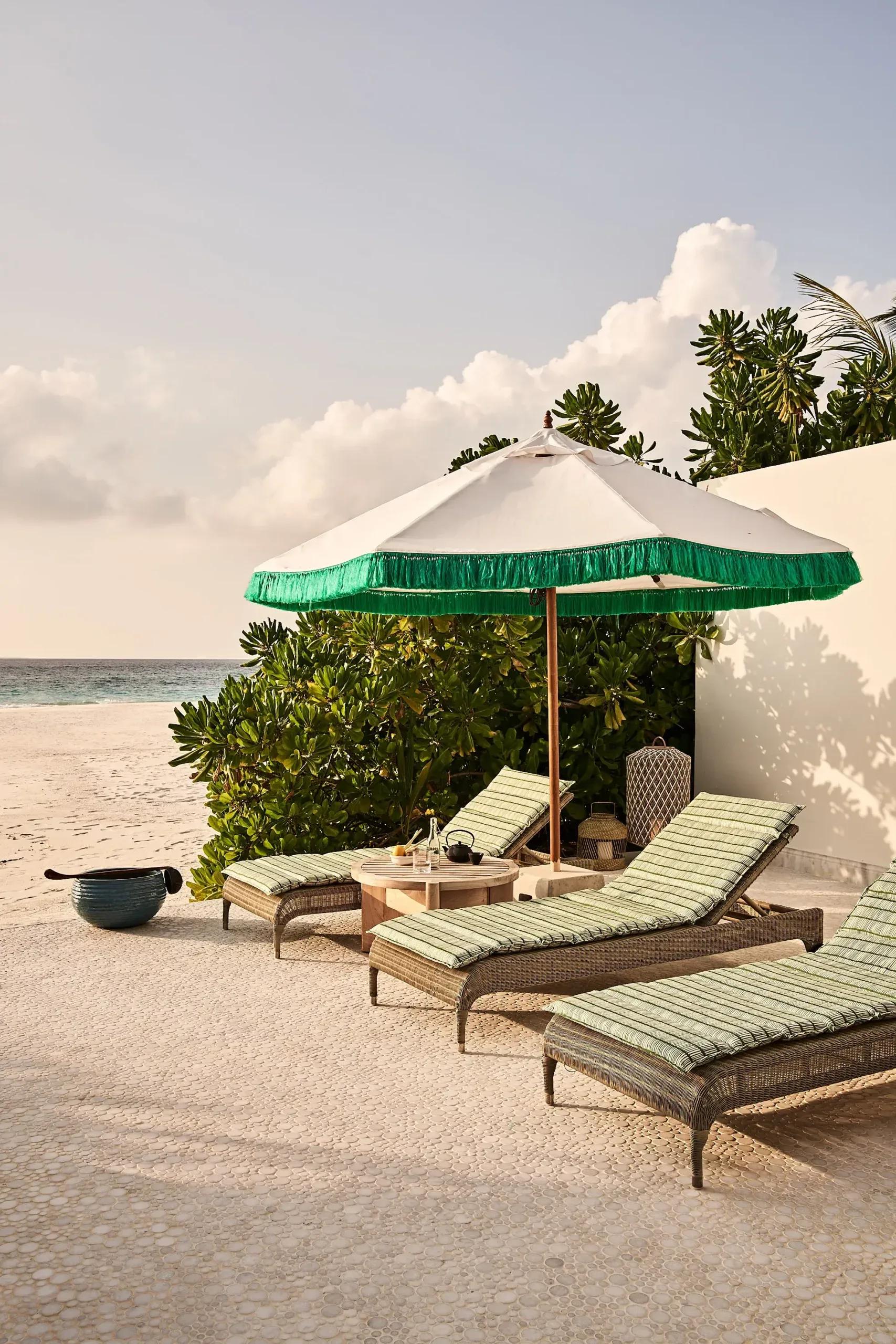 Cushioned daybeds beneath a green-fringed umbrella on Fehi Spa's sandy terrace, with the sea visible beyond a hedge
