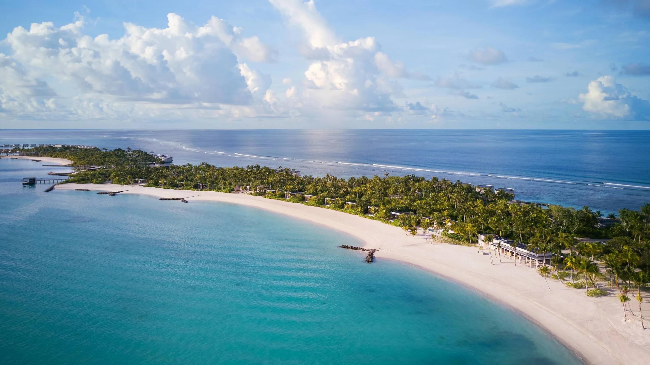 The resort's beach and reef line seen from above, palm-lined sand curving toward overwater villas at the point