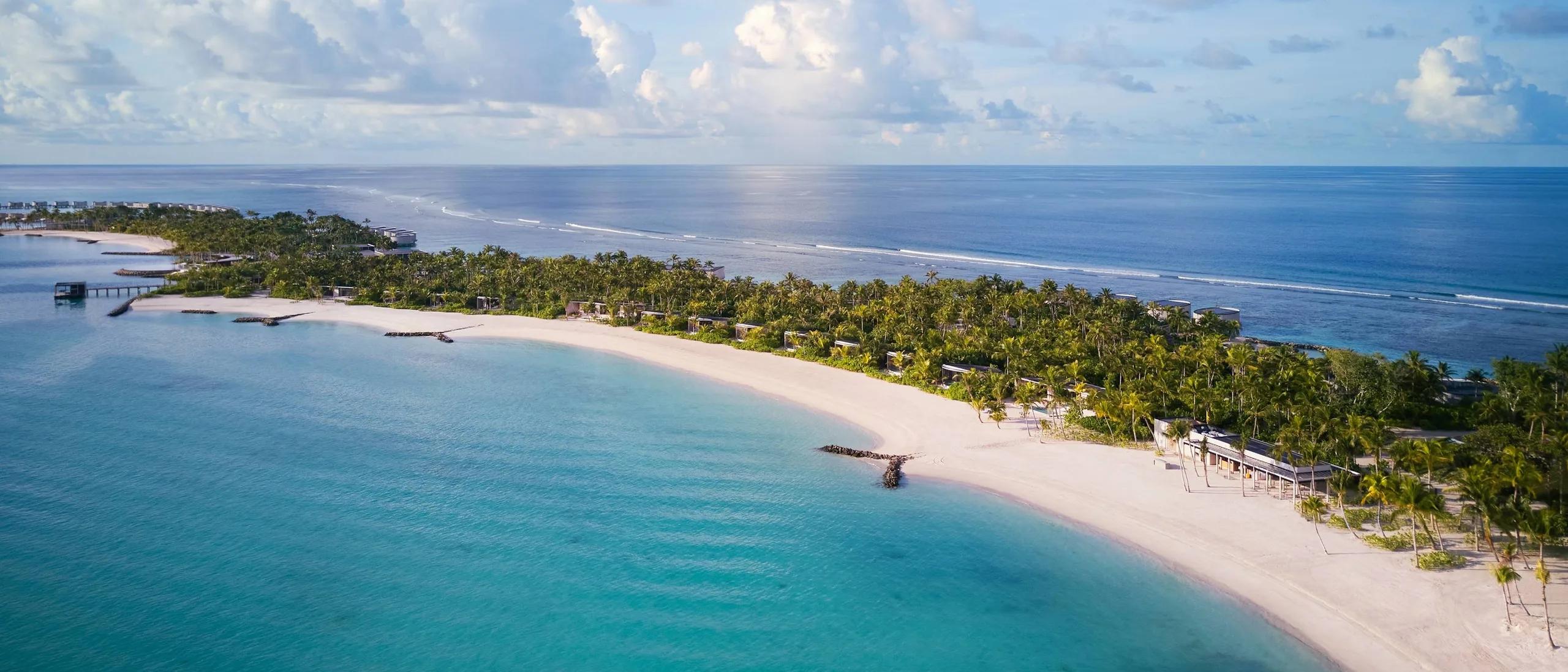 The resort's beach and reef line seen from above, palm-lined sand curving toward overwater villas at the point