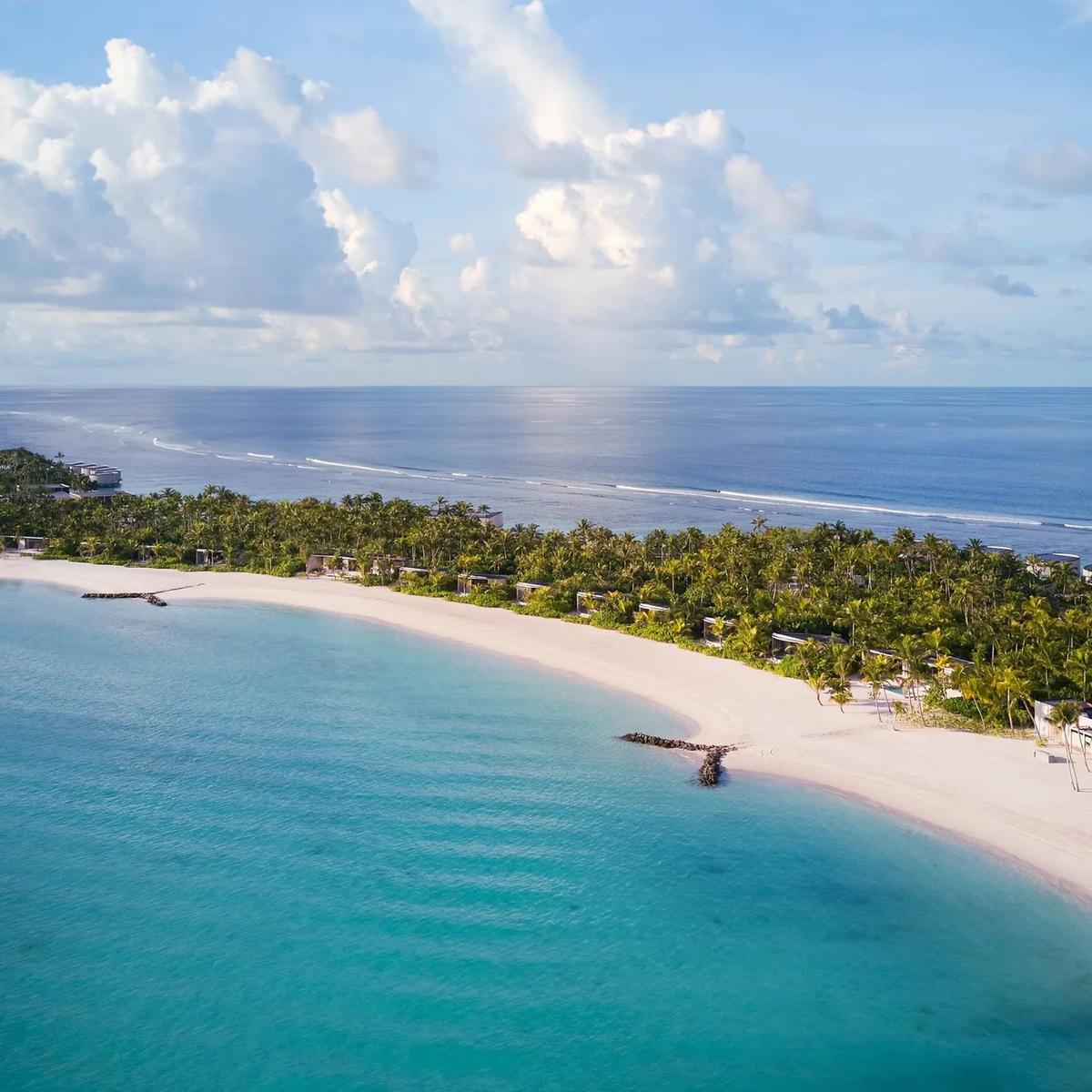 The resort's beach and reef line seen from above, palm-lined sand curving toward overwater villas at the point