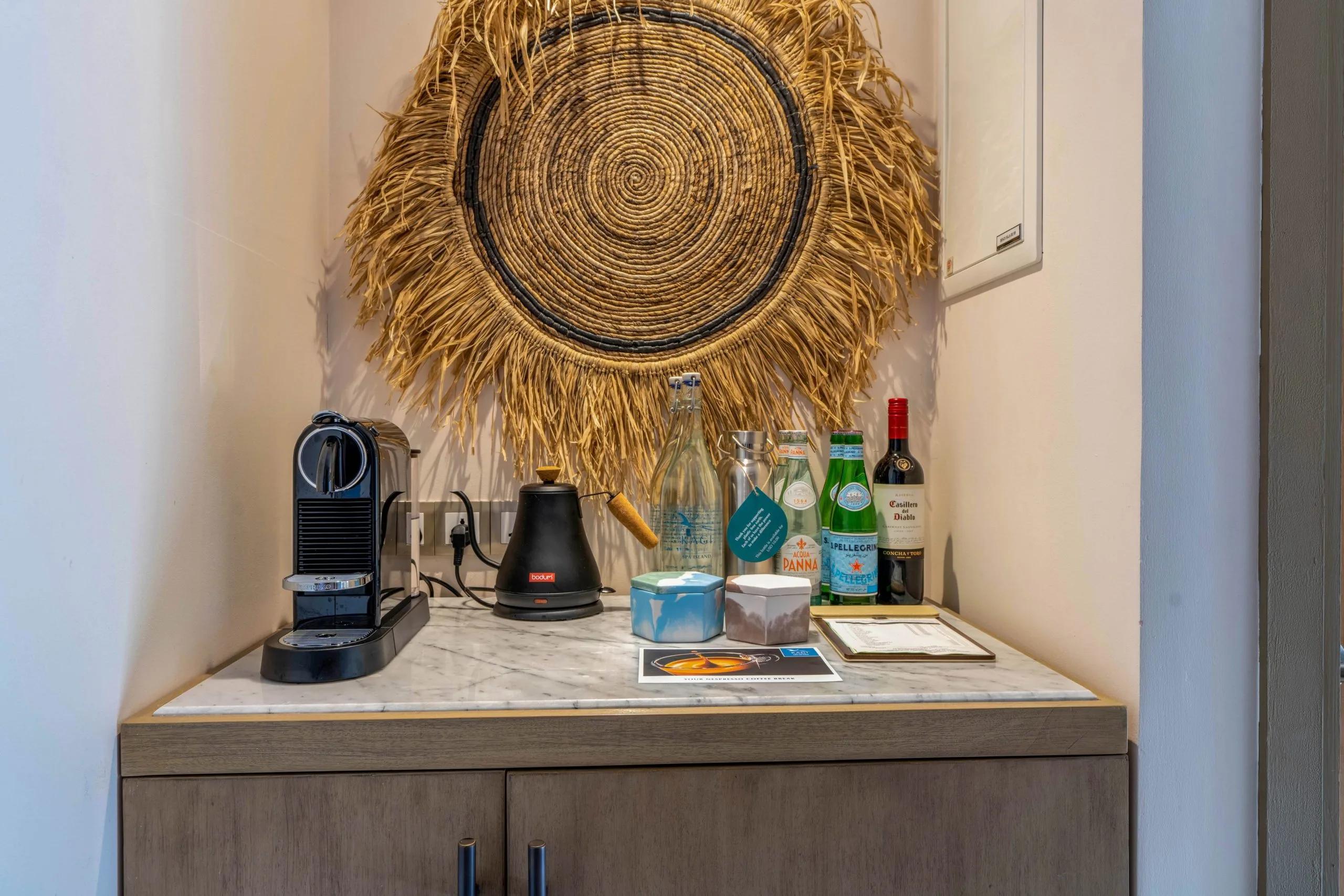 A villa's in-room bar nook with a Nespresso machine, kettle, bottled water and a bottle of red wine beneath a woven straw hanging