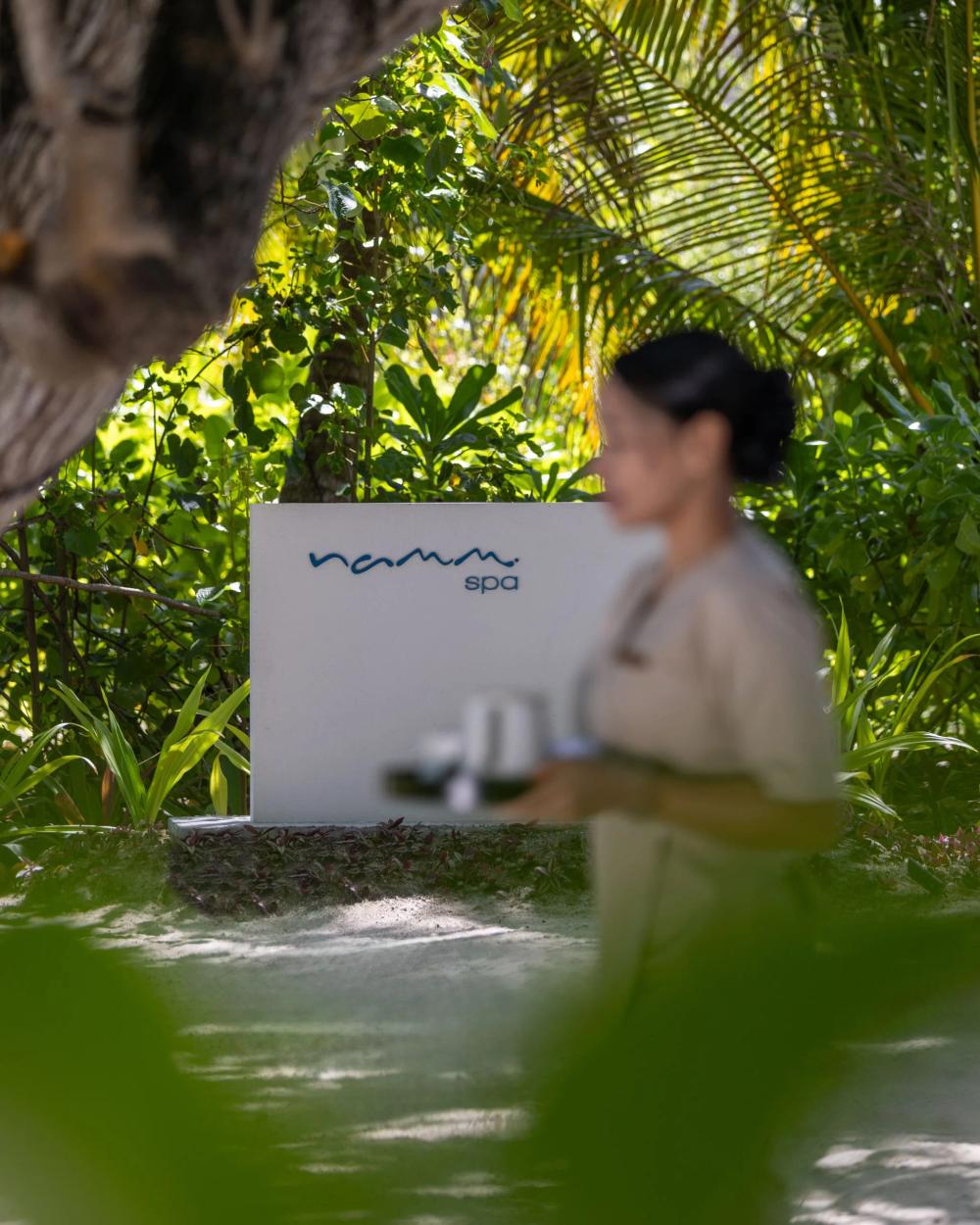 The garden path to Namm Spa at dusitD2 Feydhoo Maldives, with a therapist crossing to a treatment room