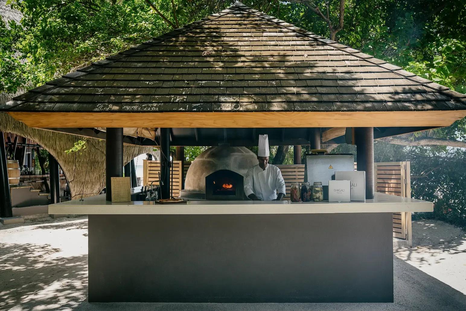 A chef sliding a pizza into the wood-fired oven at the Pizzeria, framed by its pyramid thatched roof