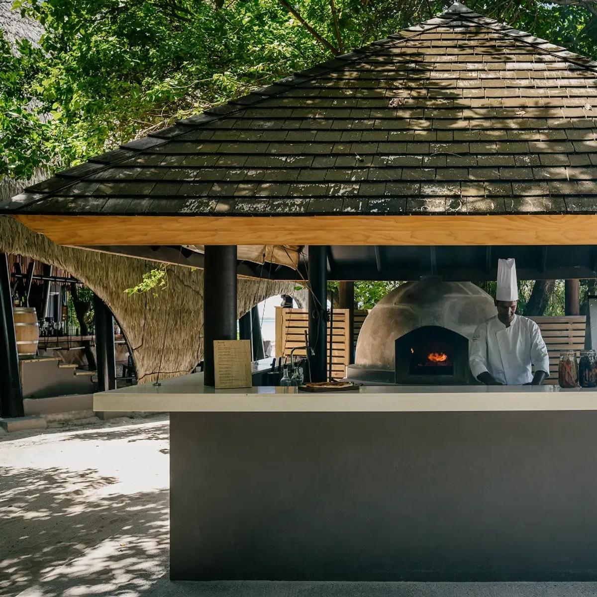 A chef sliding a pizza into the wood-fired oven at the Pizzeria, framed by its pyramid thatched roof