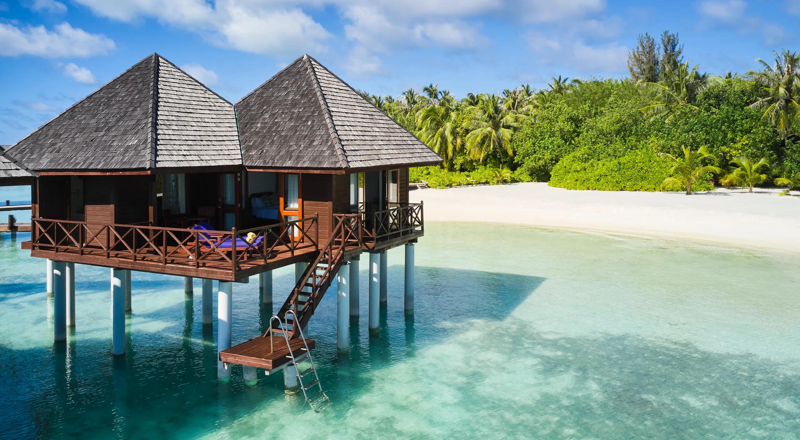 Two overwater Water Villas on stilts above a shallow turquoise lagoon, a ladder descending into the water, with palm trees and beach behind