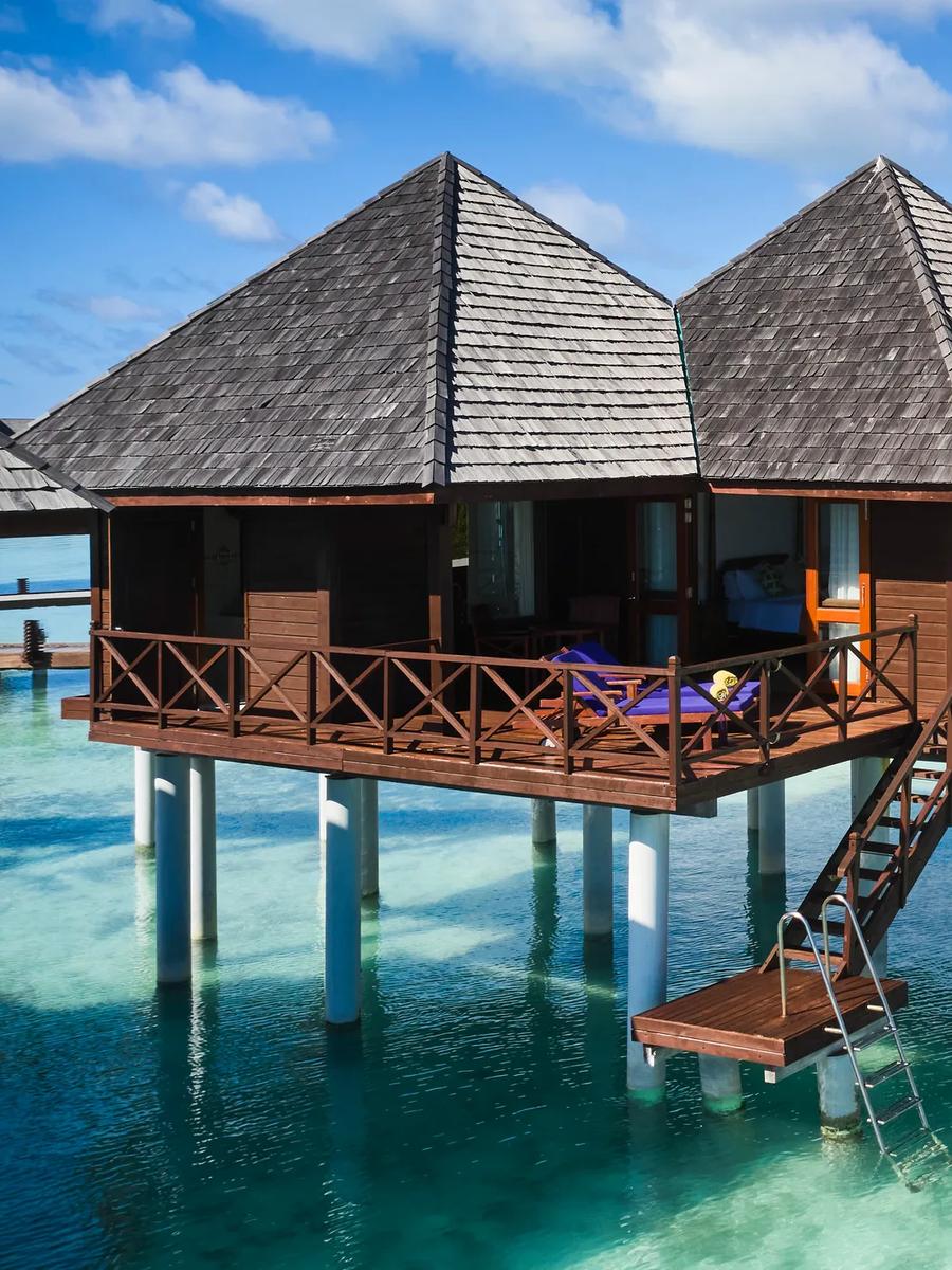 Two overwater Water Villas on stilts above a shallow turquoise lagoon, a ladder descending into the water, with palm trees and beach behind
