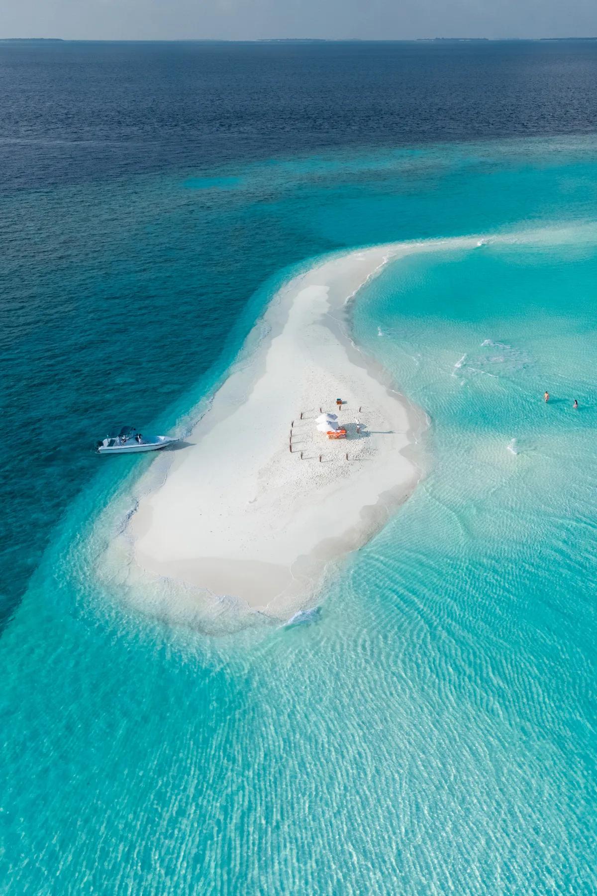 An aerial view of a private picnic set up on a bare sandbank, a boat moored alongside and two guests wading in the shallows nearby