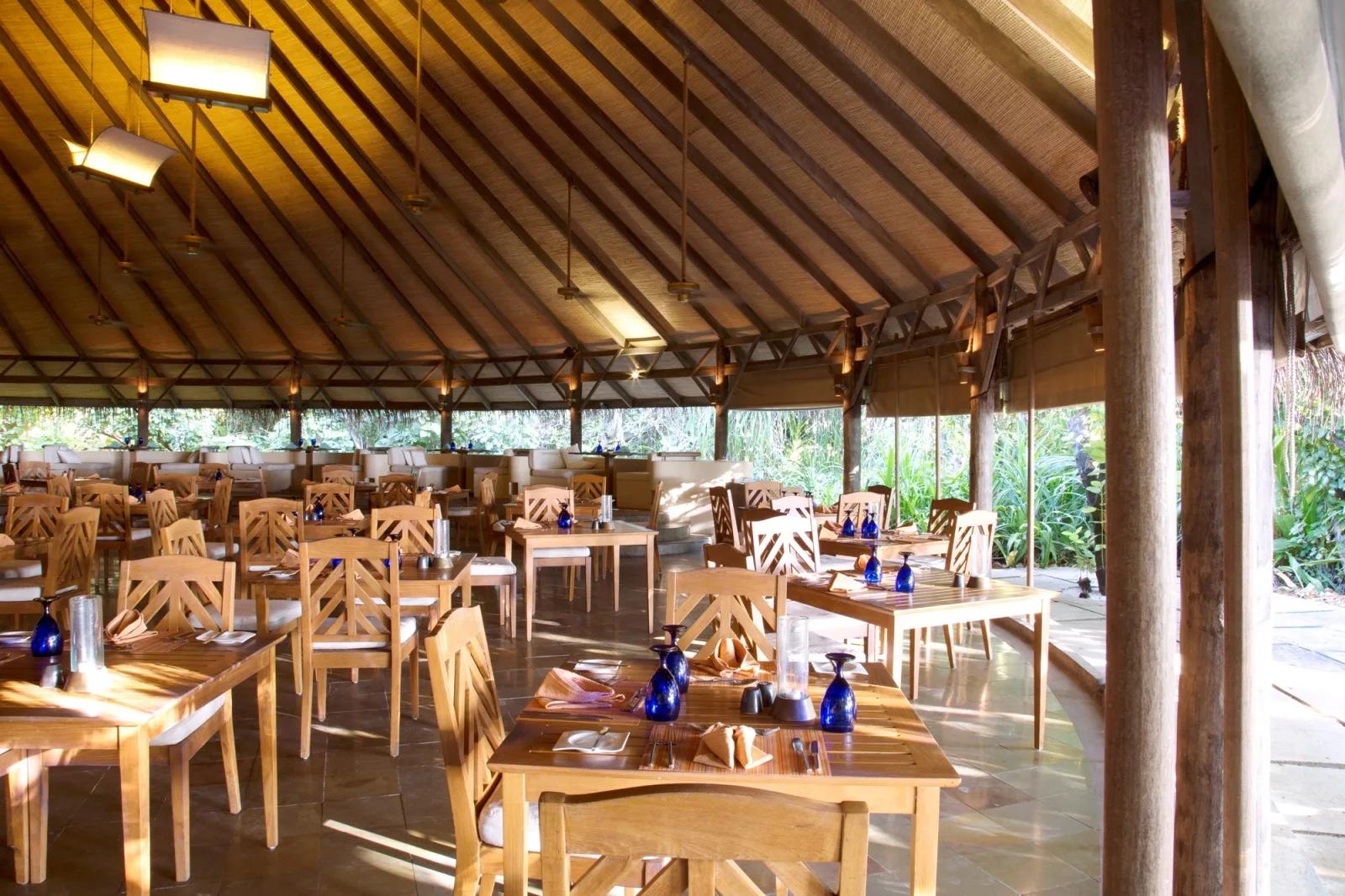 Cowrie restaurant's dining room set for service under a high thatched roof, blue glassware on the tables