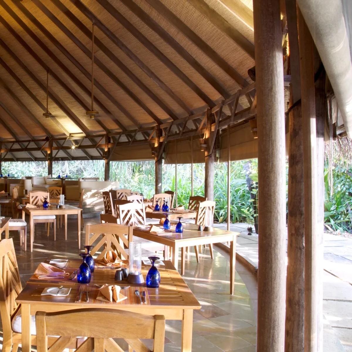 Cowrie restaurant's dining room set for service under a high thatched roof, blue glassware on the tables