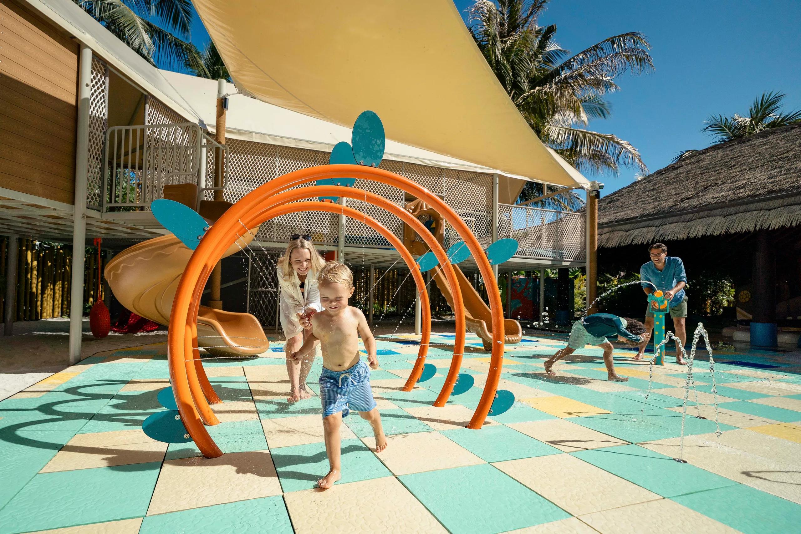 Children run through a water-spraying arch structure at Lha Velaa Kids Club, a slide and play tower behind them