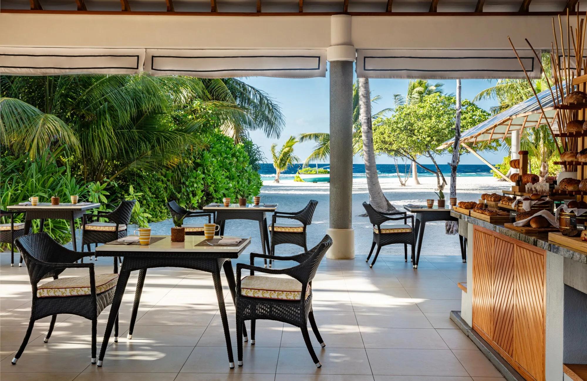 REEF restaurant's open-air breakfast terrace, wicker chairs at set tables beneath a thatched roof opening onto a white-sand beach and the lagoon beyond