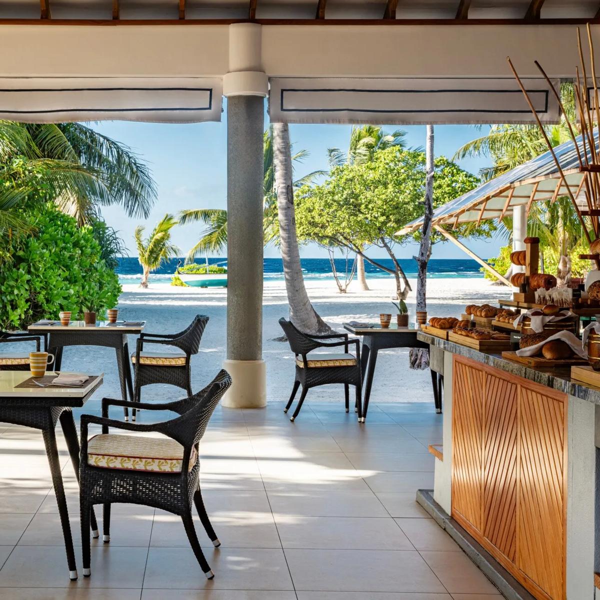 REEF restaurant's open-air breakfast terrace, wicker chairs at set tables beneath a thatched roof opening onto a white-sand beach and the lagoon beyond