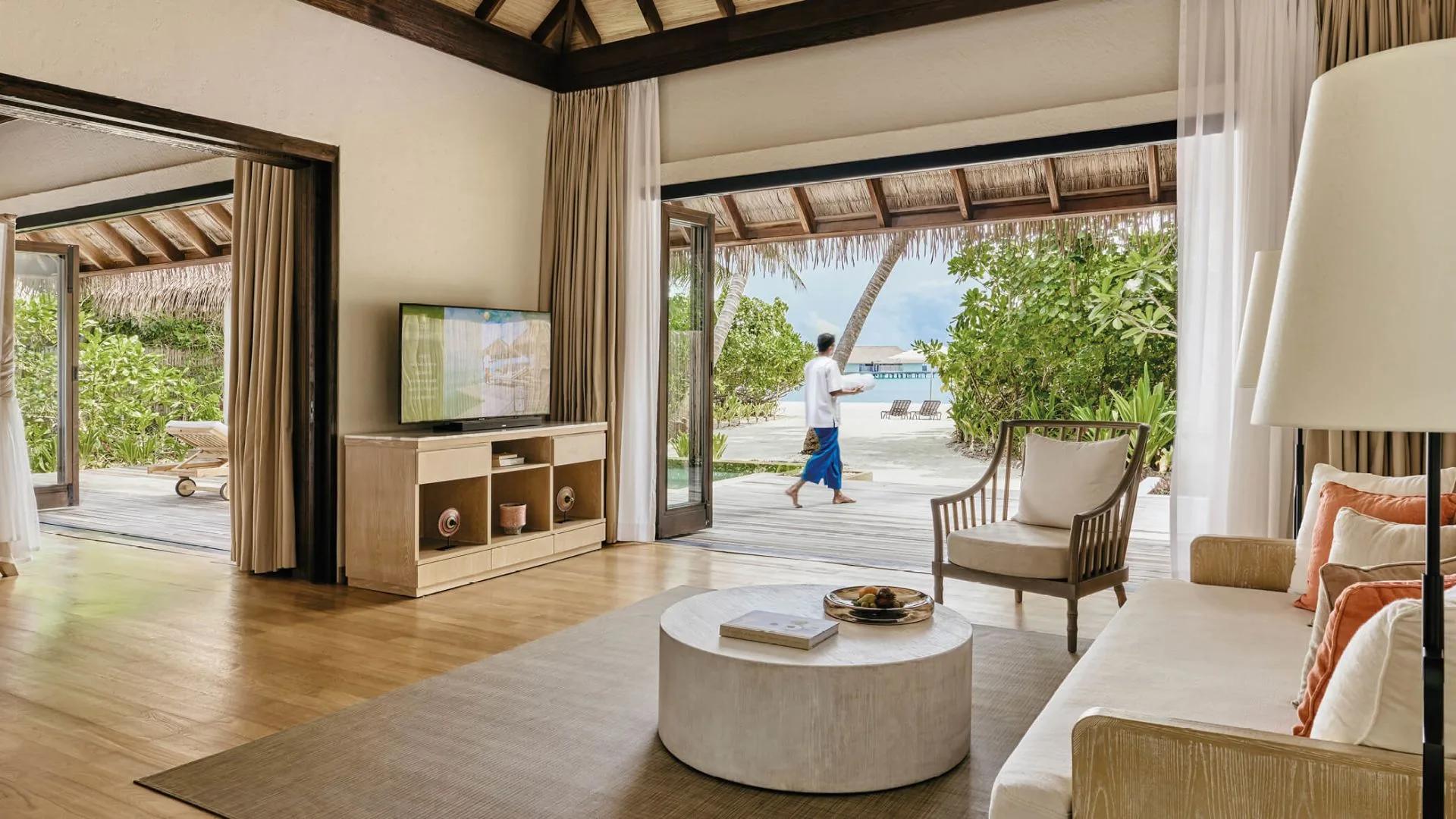 Villa living room with white sofas and a television, staff member carrying a tray past open doors onto a beachfront deck