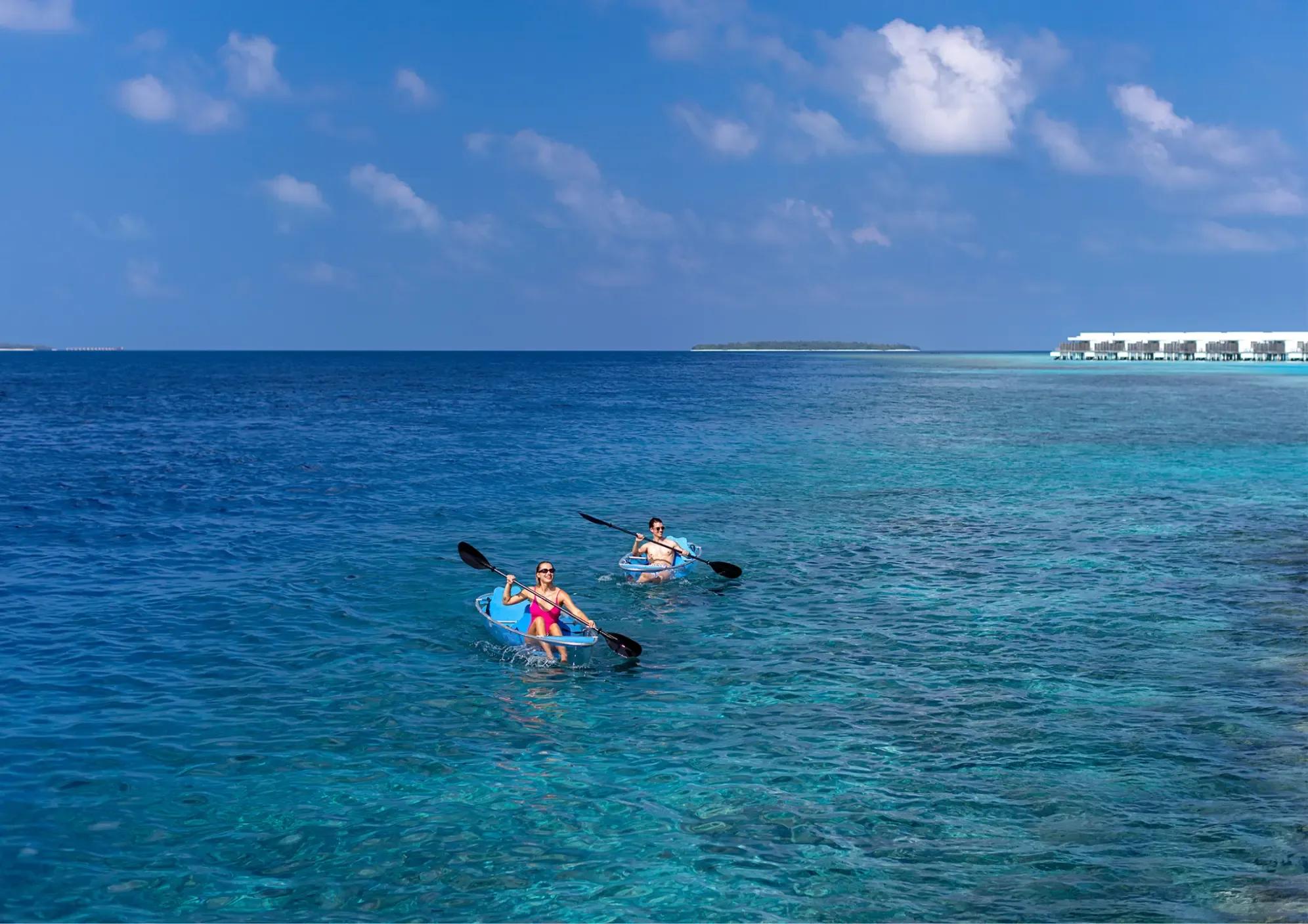 Two guests paddling clear-bottom kayaks on the lagoon, Niva Dhigali's overwater villas in the distance