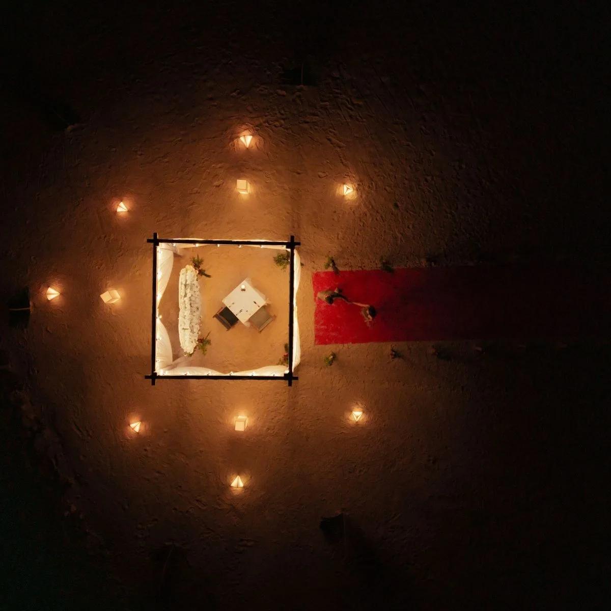 Aerial view of a beach wedding ceremony set up at SO/ Maldives at dusk, a square canopy and red aisle ringed by lanterns on the sand