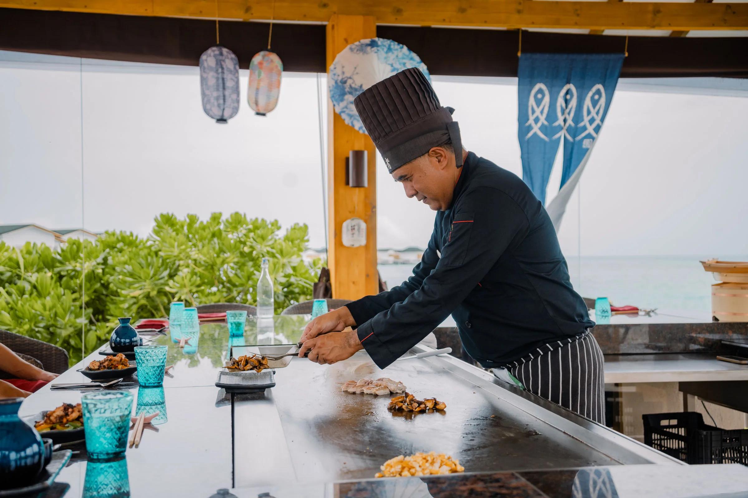A chef cooks on the teppanyaki hot plate at Japanese Corner, lagoon views visible through the open-sided dining room