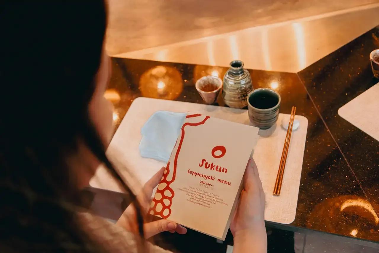 A guest reading Sukun's teppanyaki menu card at the table, sake service laid out alongside