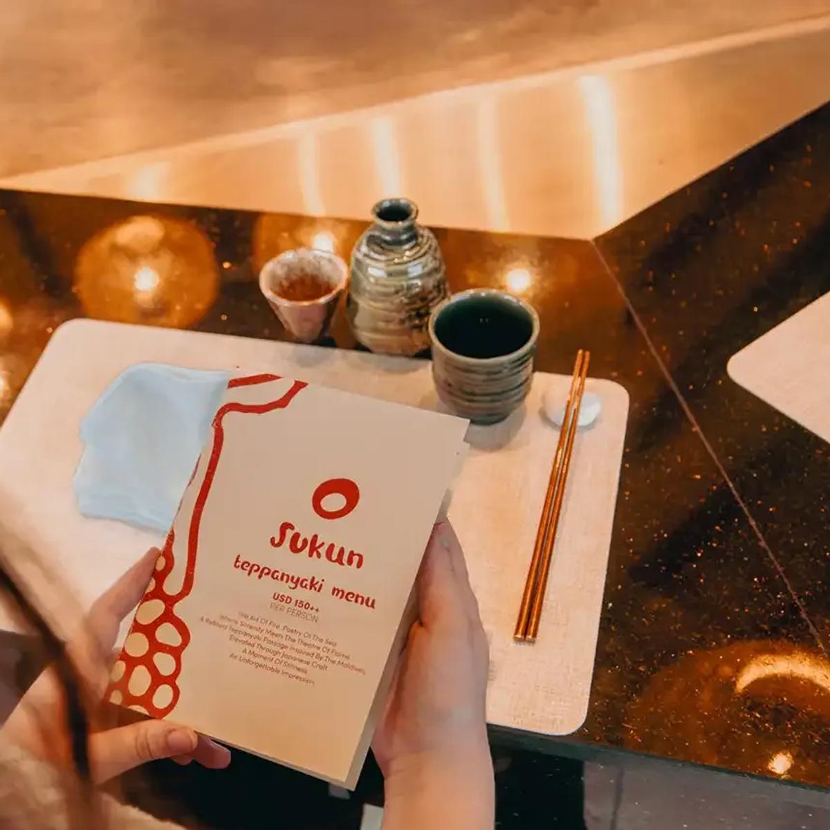 A guest reading Sukun's teppanyaki menu card at the table, sake service laid out alongside