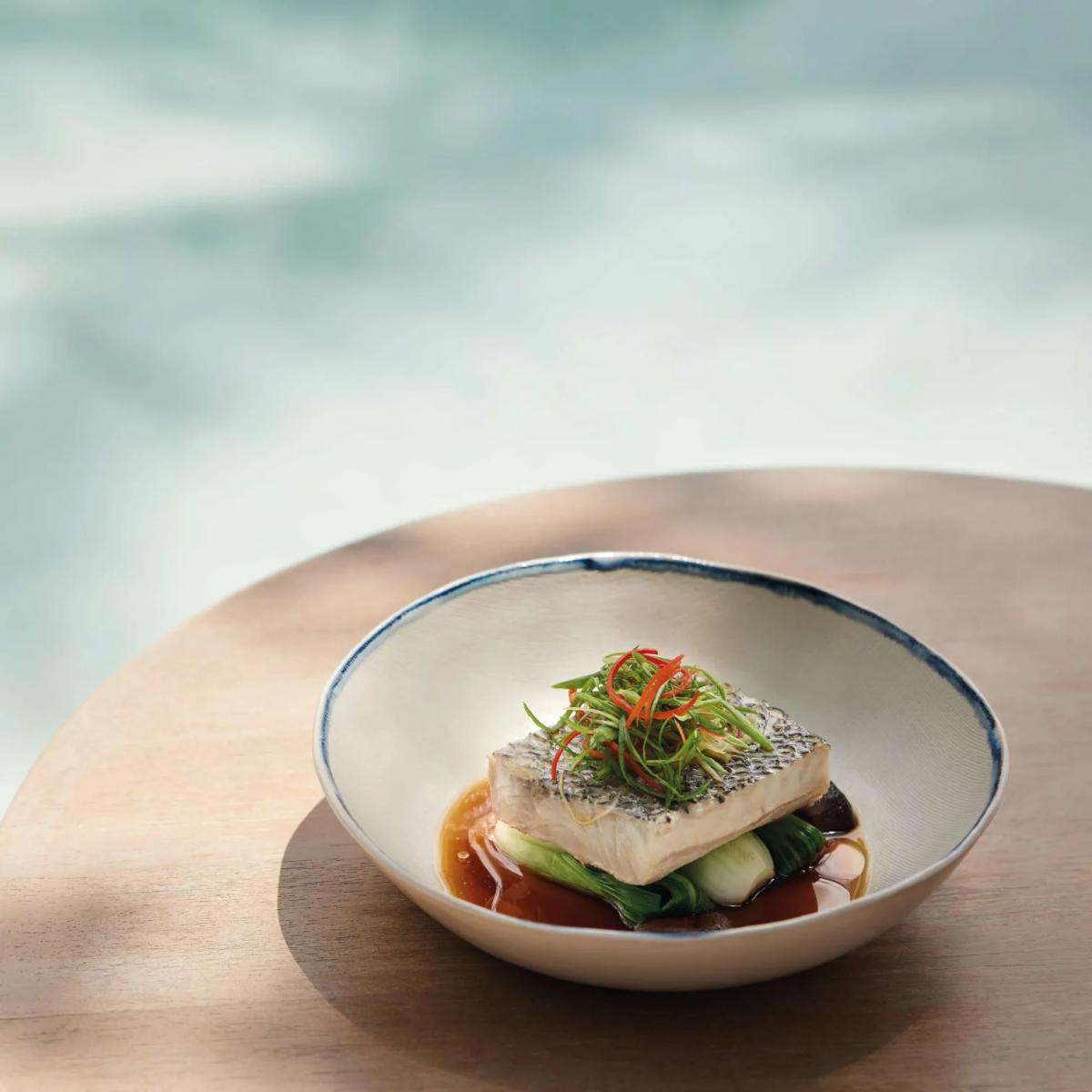 Plated fish dish with pickled vegetables and chilli garnish on a ceramic plate, lagoon visible in the background