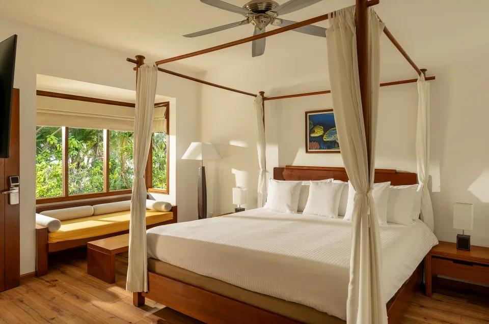 Beach Bungalow bedroom with a four-poster canopy bed, white bedding, and a daybed with a mustard-yellow cushion beneath a garden-facing window