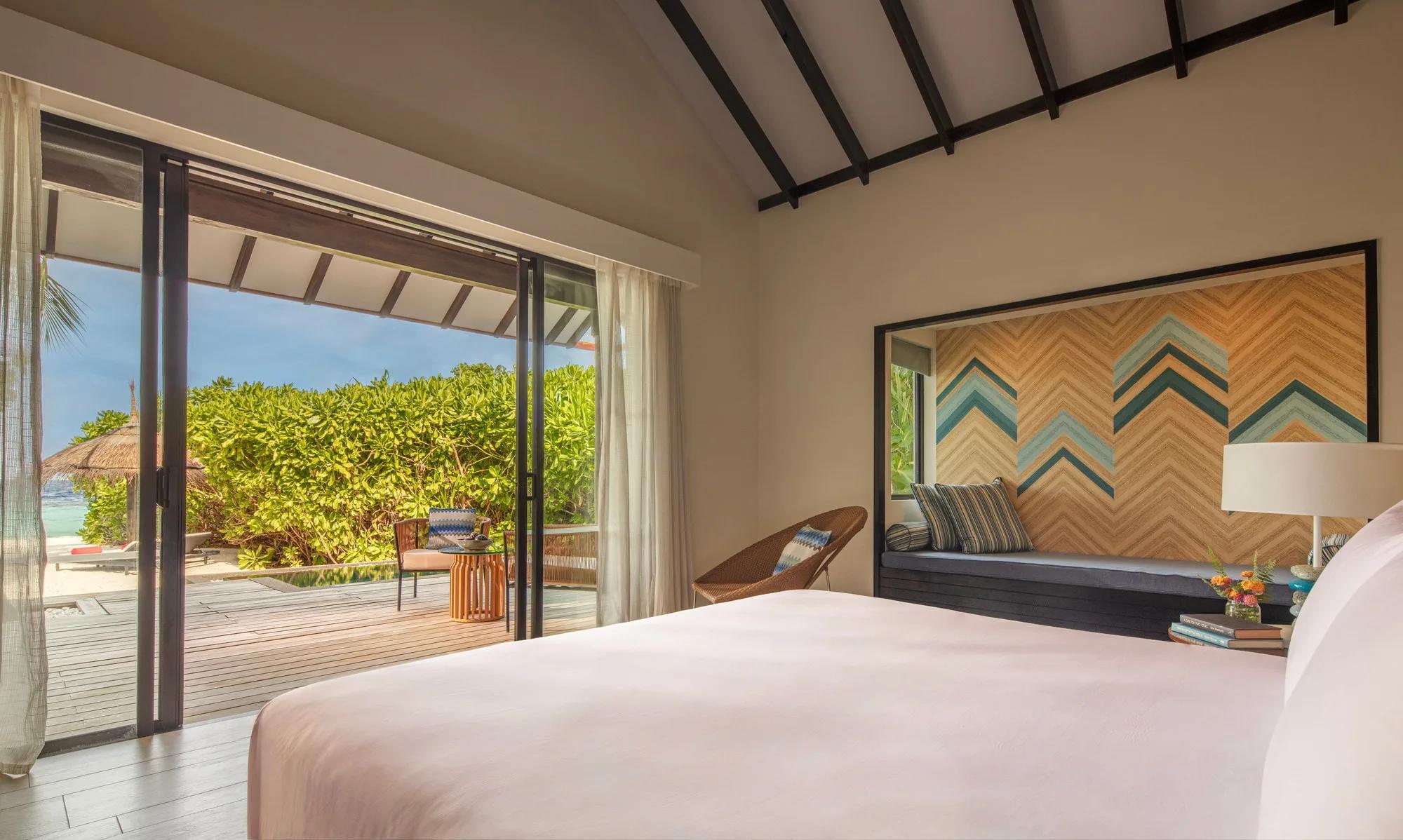 A Sunset Beach Pool Villa's bed facing an open deck, a dense green hedge and the private pool's edge visible beyond the sliding doors