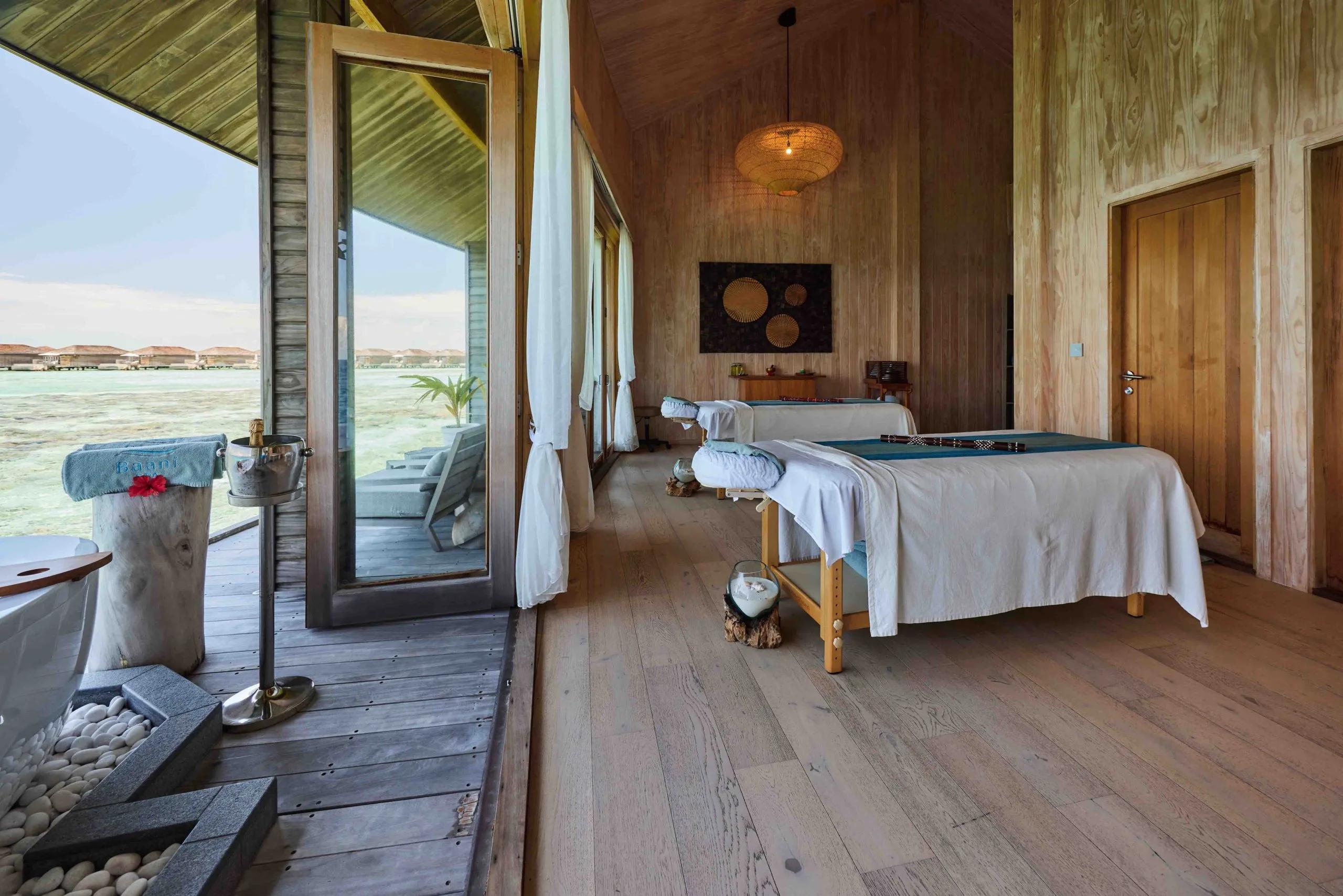 An overwater spa treatment room with two massage beds, sliding doors open to the ocean and a freestanding bathtub on the deck outside
