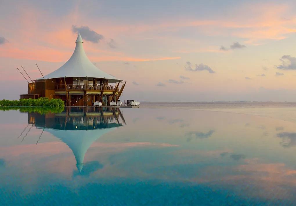Wide view of Baros's cone-roofed overwater dining pavilion at dusk, its silhouette reflected in the still lagoon with palms along the shoreline