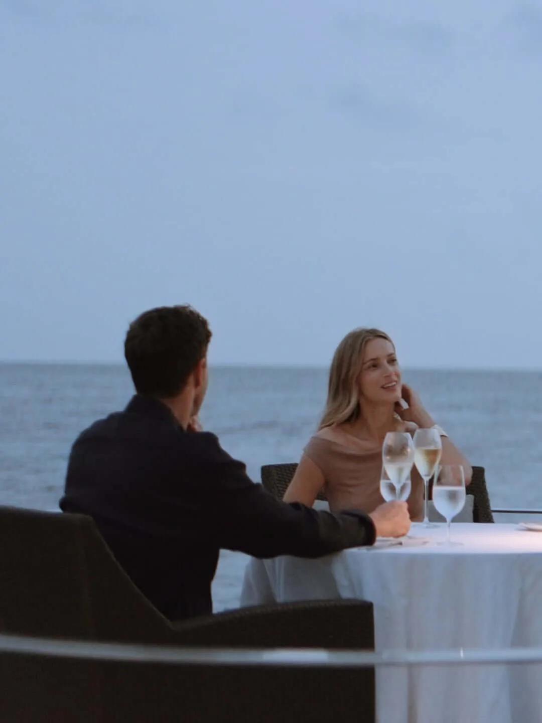 A couple share a candlelit table beside the ocean at dusk, glasses of champagne poured