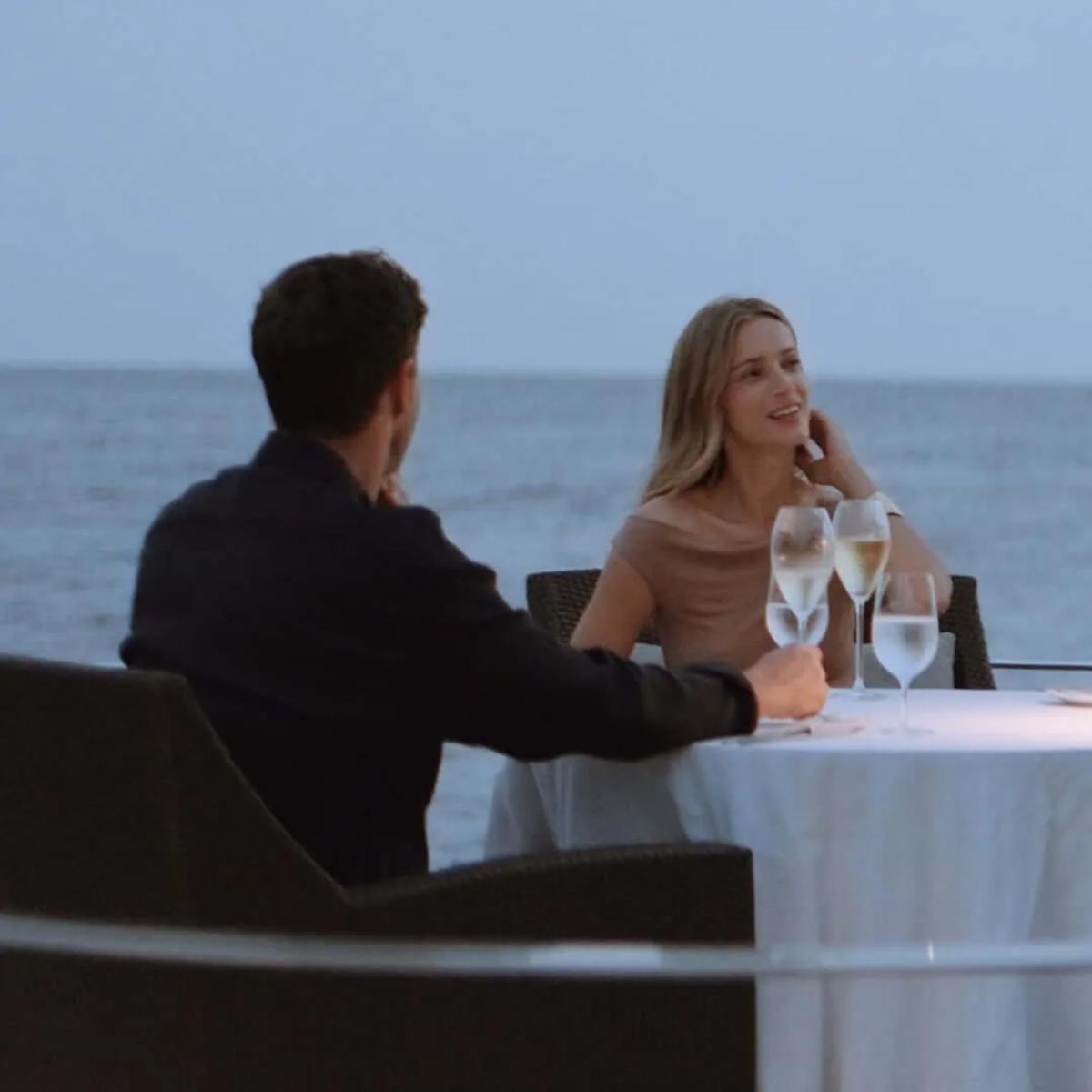 A couple share a candlelit table beside the ocean at dusk, glasses of champagne poured
