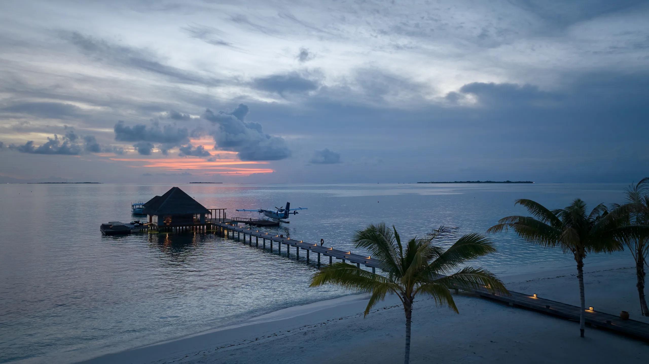 A seaplane moored at the overwater lounge pontoon at dusk, palm trees silhouetted against a pink and blue sky.