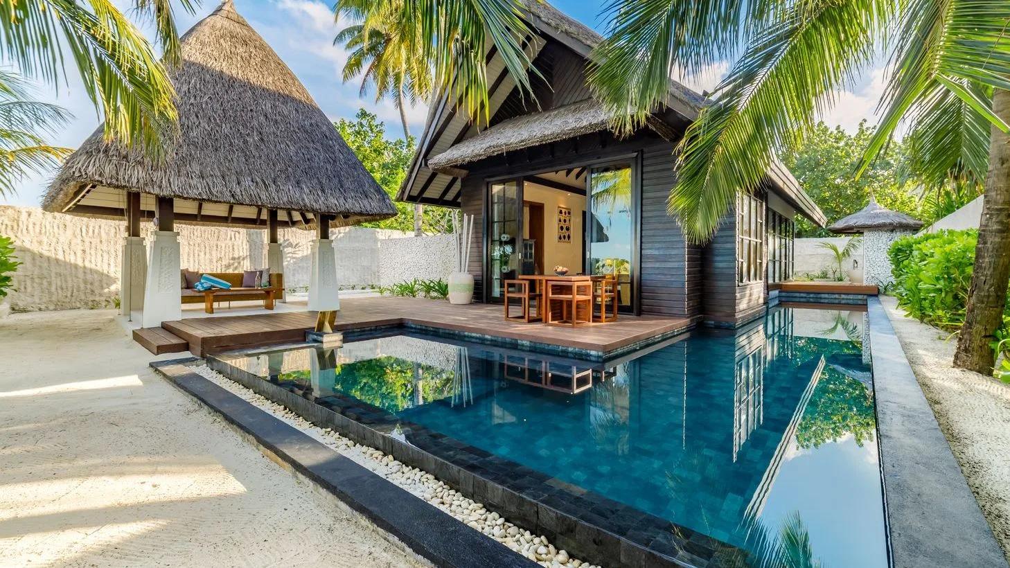 An Earth Pool Villa's private L-shaped pool and thatched daybed pavilion, framed by palm trees on the beach