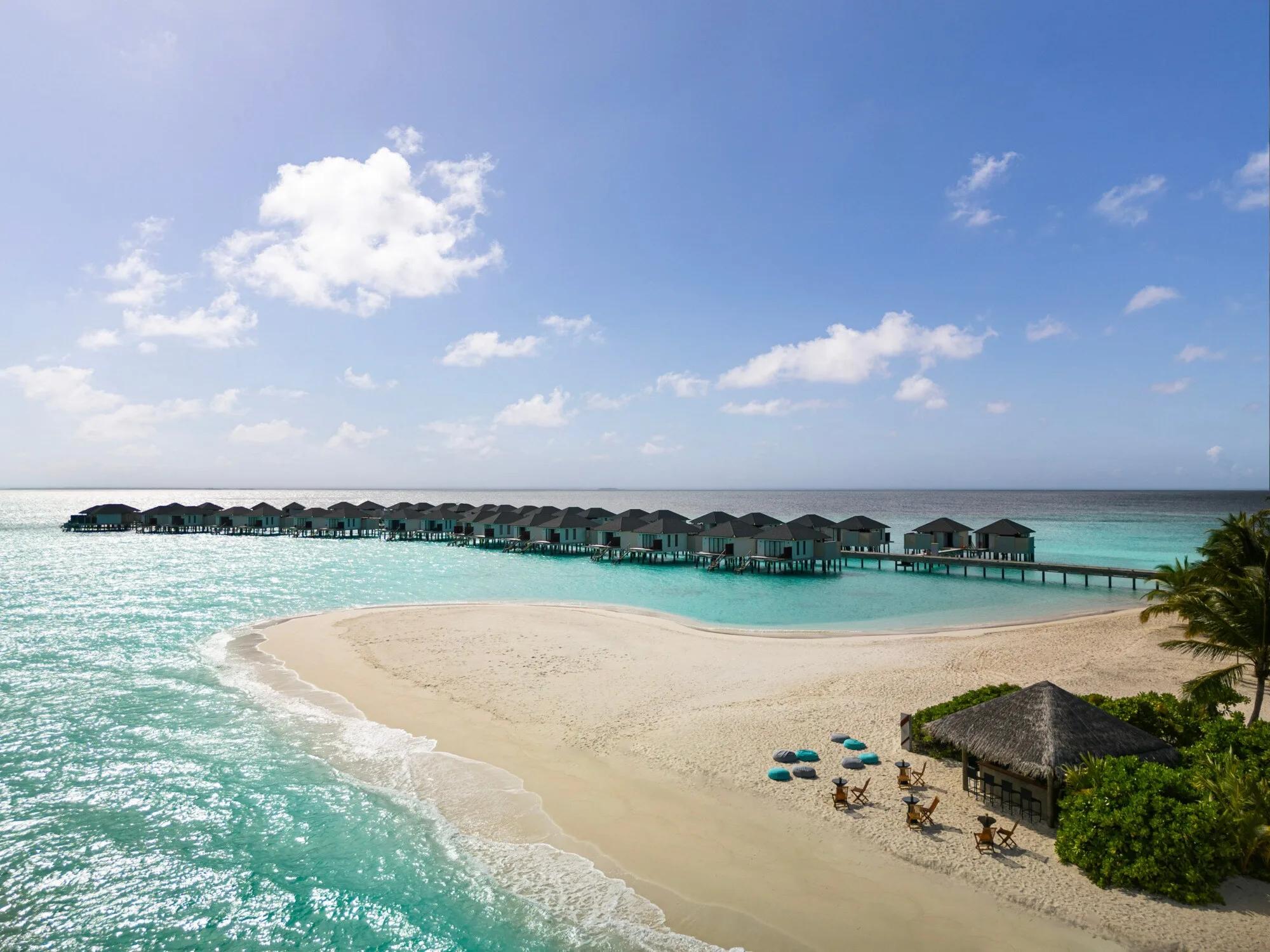 Aerial view of a row of overwater villas on stilts leading out from a white sandbank, a thatched beach bar and loungers set at the sand's edge