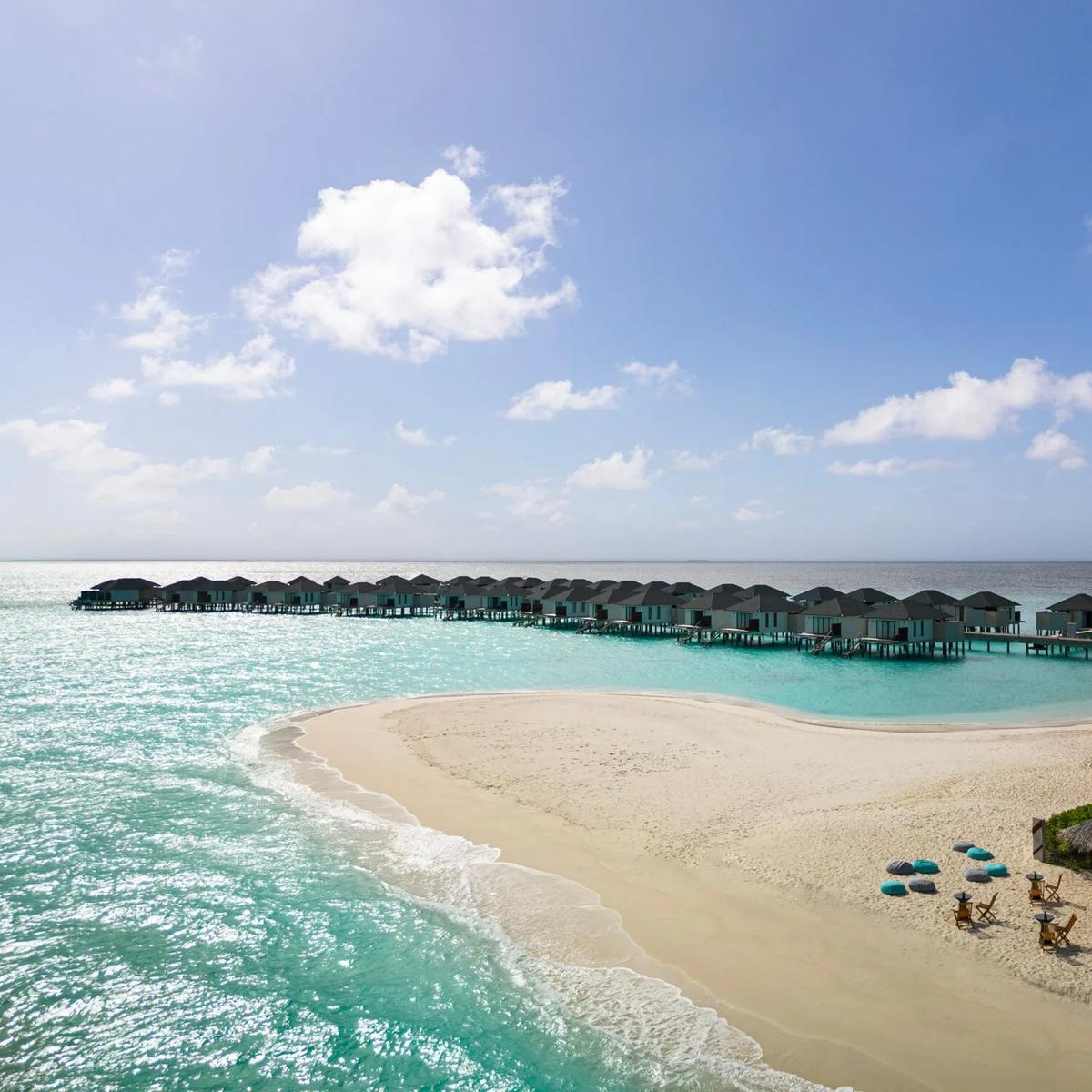 Aerial view of a row of overwater villas on stilts leading out from a white sandbank, a thatched beach bar and loungers set at the sand's edge