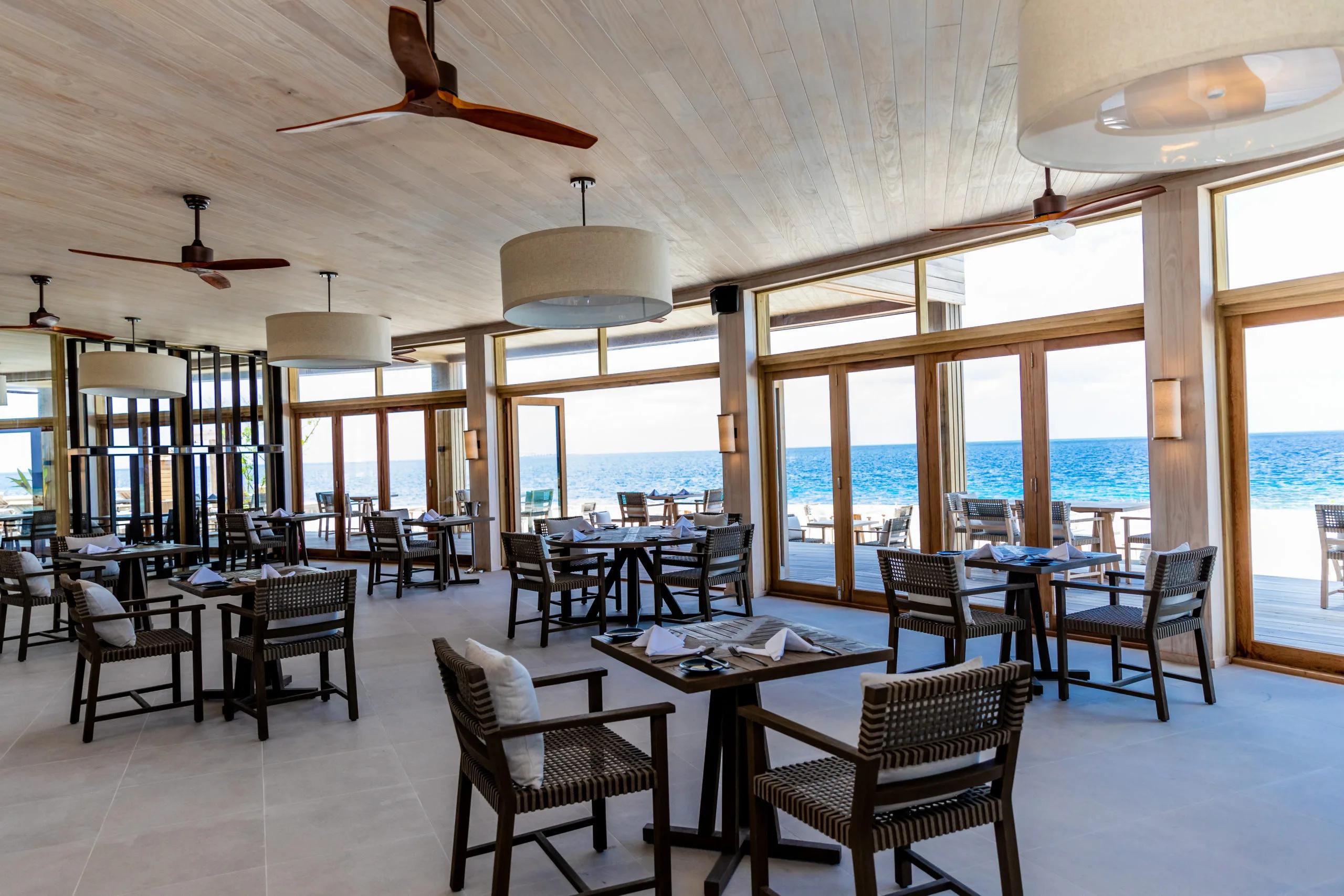 Noo Faru's dining room, tables set beneath ceiling fans and pendant lights, floor-to-ceiling glass doors open to the ocean