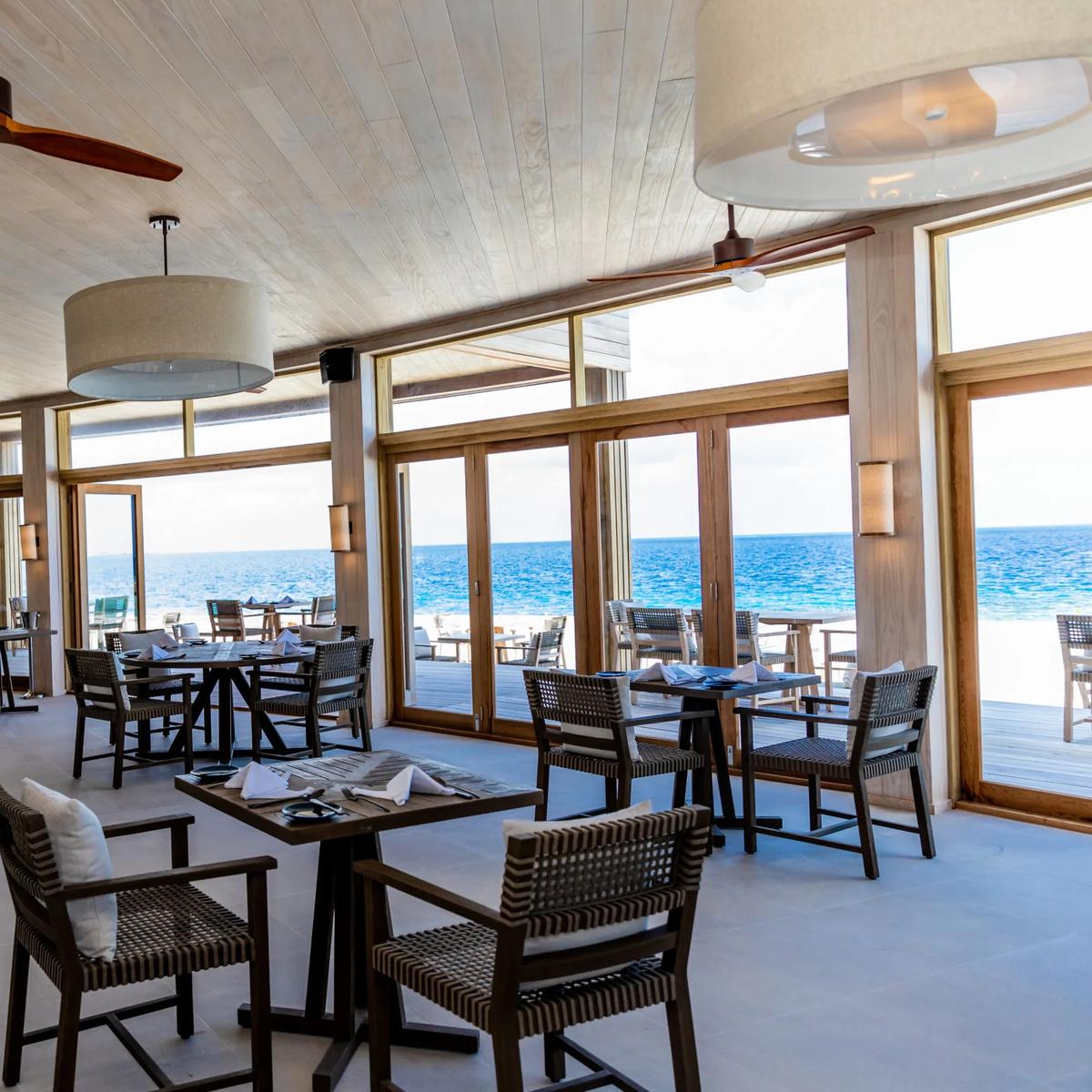 Noo Faru's dining room, tables set beneath ceiling fans and pendant lights, floor-to-ceiling glass doors open to the ocean