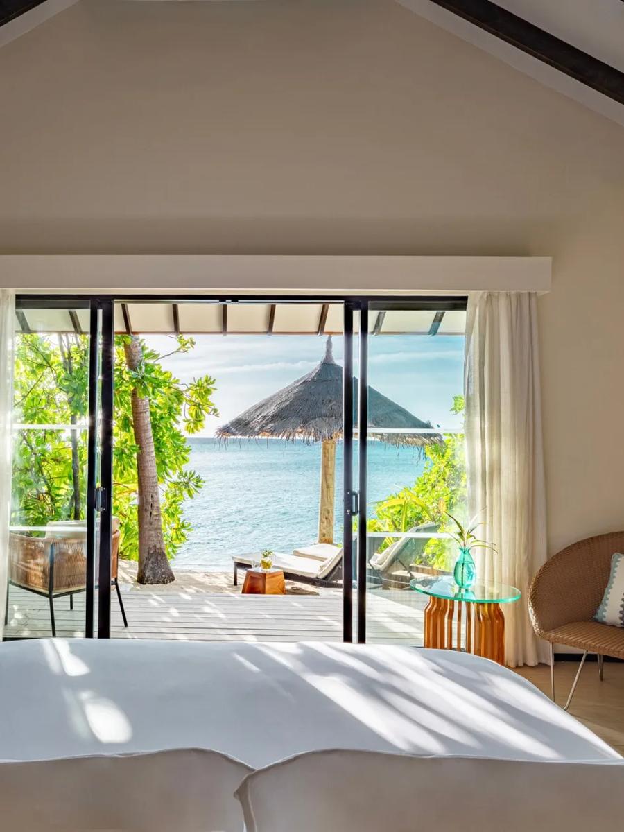 A view from a Beach Villa with Private Pool's bed out through sliding doors to a thatched beach umbrella and the lagoon beyond