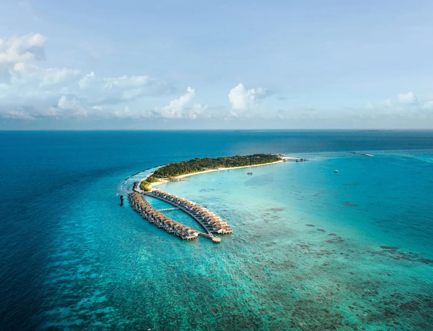 Wide aerial view of Sirru Fen Fushi's full island, its overwater villa jetty extending into the lagoon on one side and reef stretching to the horizon