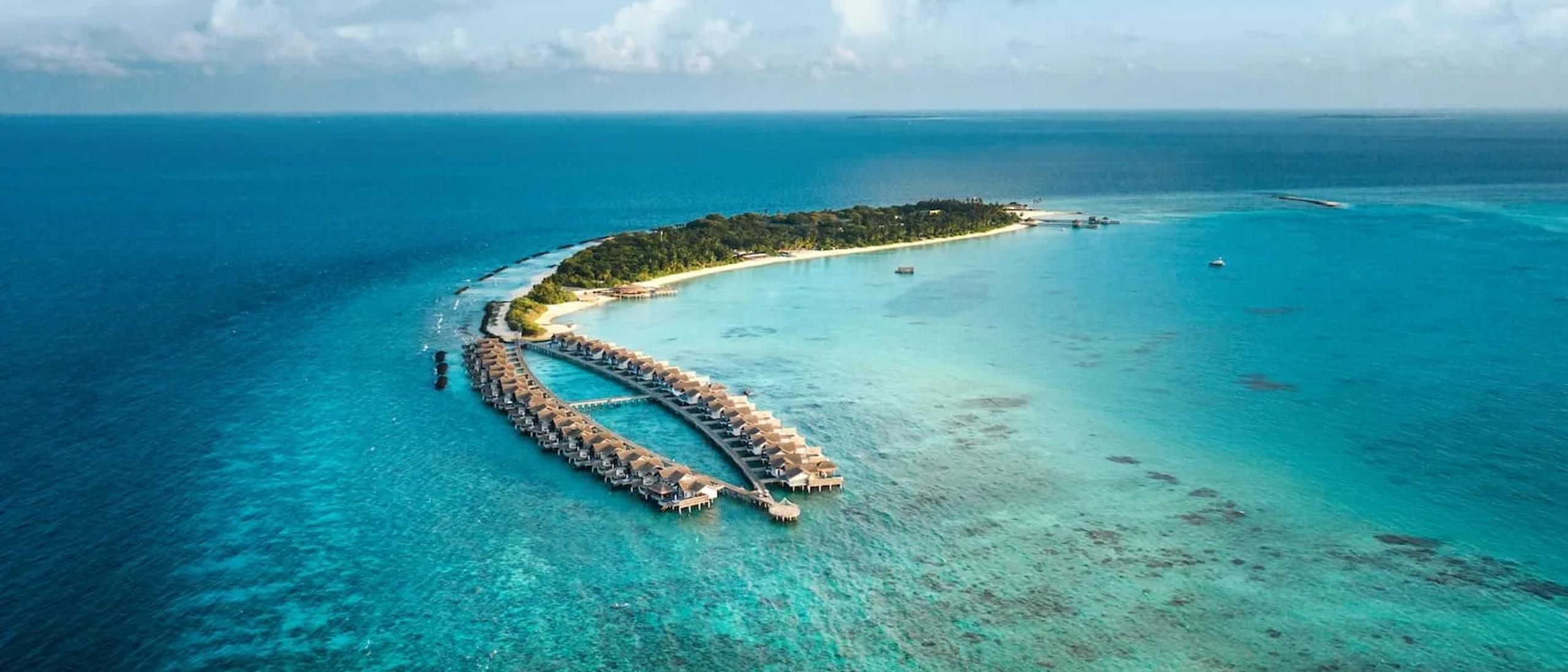 Wide aerial view of Sirru Fen Fushi's full island, its overwater villa jetty extending into the lagoon on one side and reef stretching to the horizon