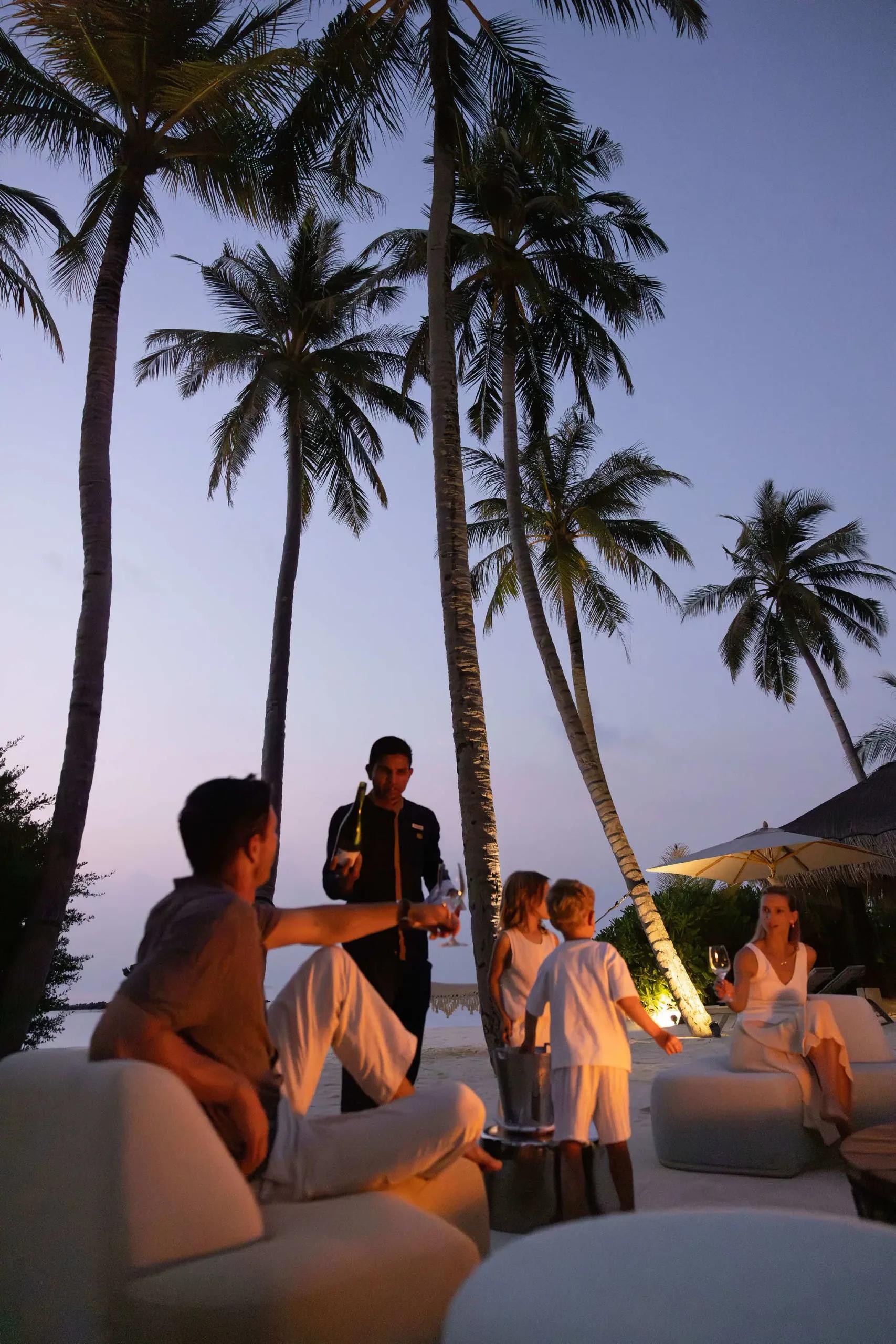 A waiter pours champagne for a family gathered on the beach under palm trees at dusk