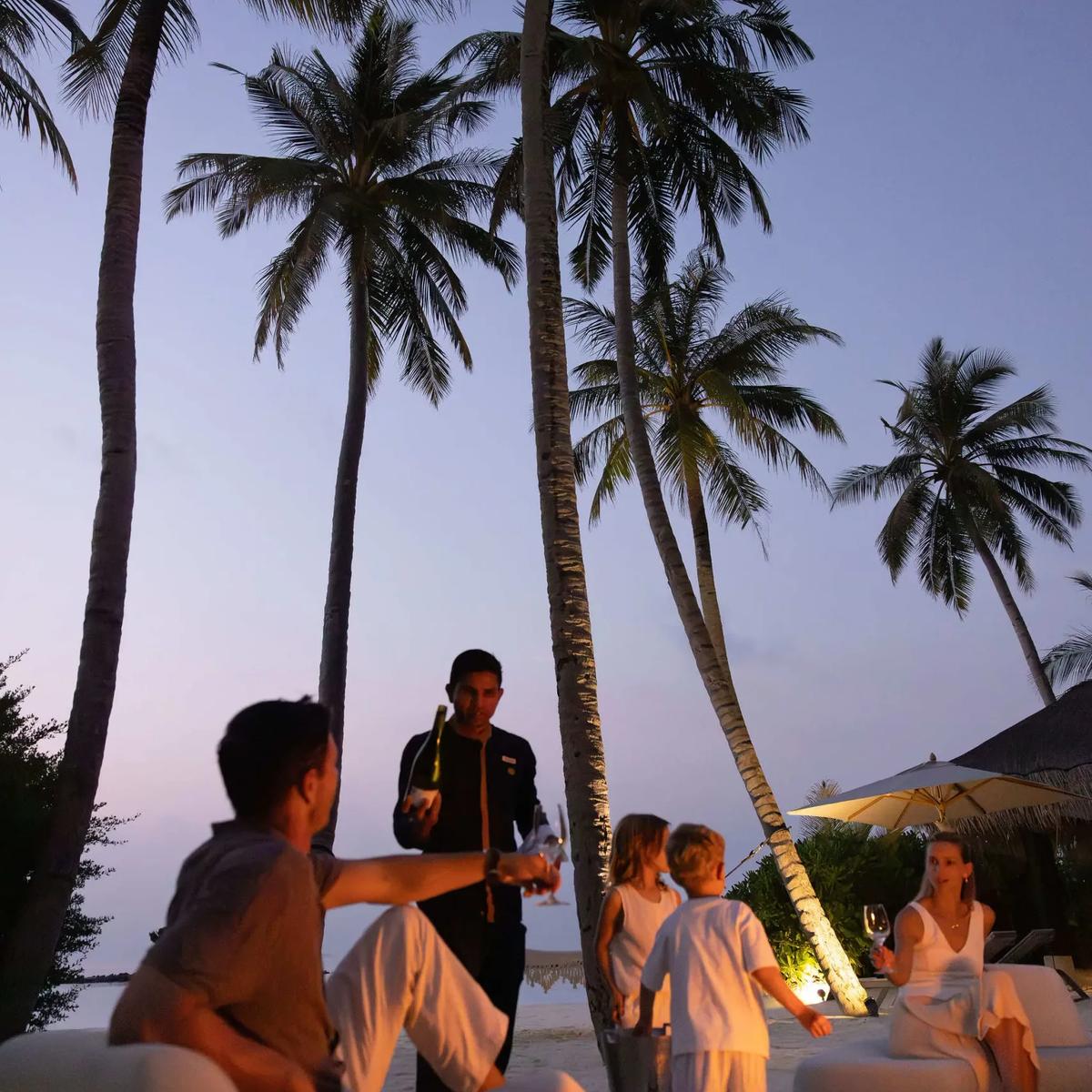 A waiter pours champagne for a family gathered on the beach under palm trees at dusk