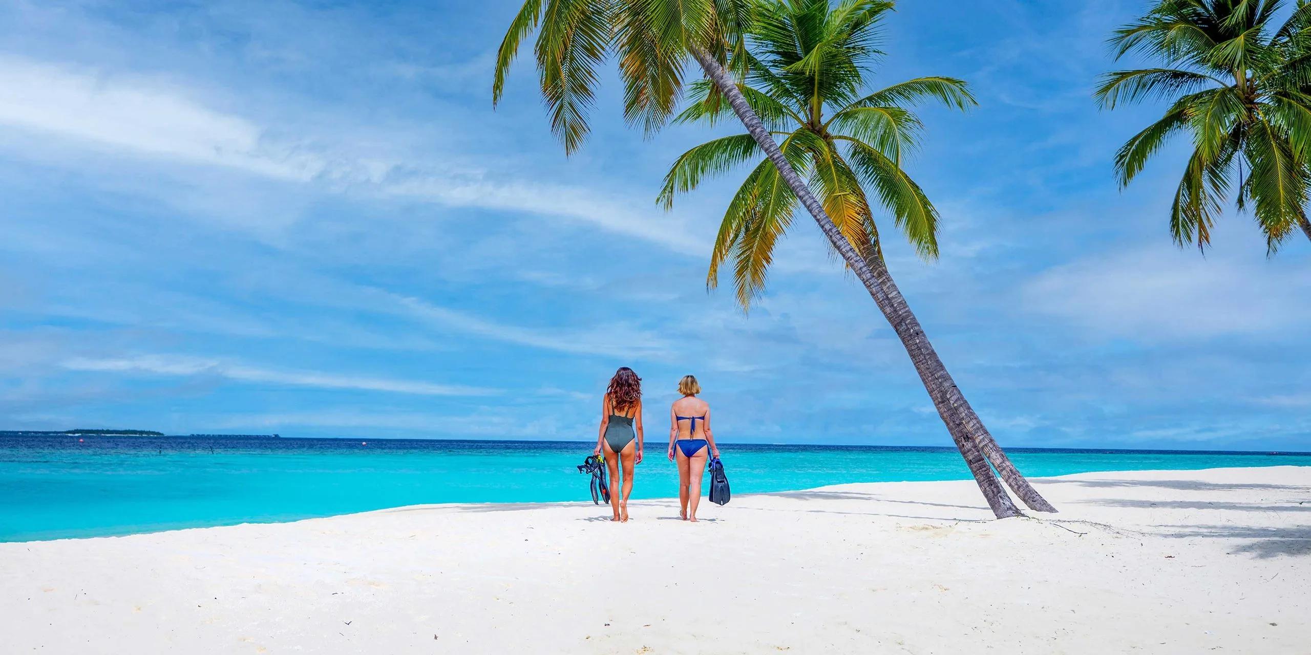 Two guests carrying snorkel fins as they walk from the beach toward Baglioni Maldives' house reef