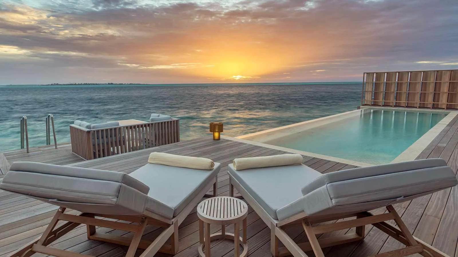 Sun loungers and an infinity pool on an overwater residence deck as the sky turns orange over a distant shoreline