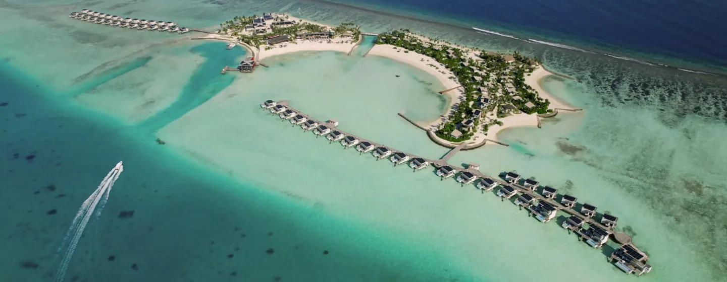 Aerial view of SO/ Maldives' island with a speedboat crossing the lagoon toward it, overwater villas lining both jetties and beach villas set among the palms