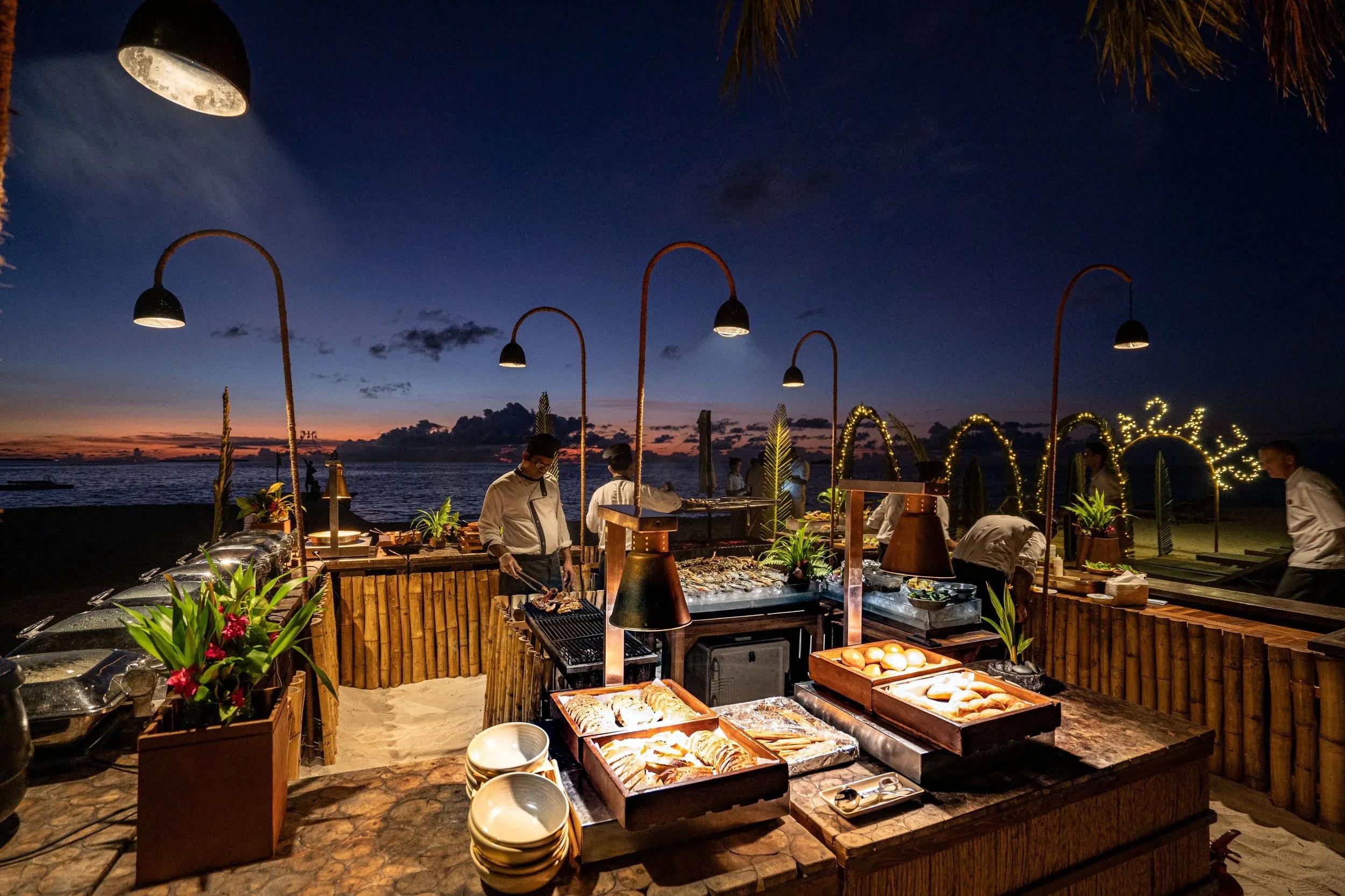 Chefs grilling seafood at Sea Salt, Ayada's beach barbecue, under strings of lamps at dusk