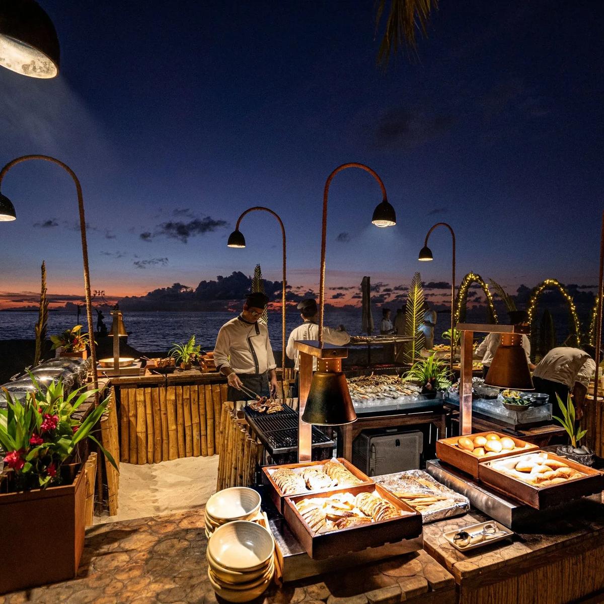 Chefs grilling seafood at Sea Salt, Ayada's beach barbecue, under strings of lamps at dusk