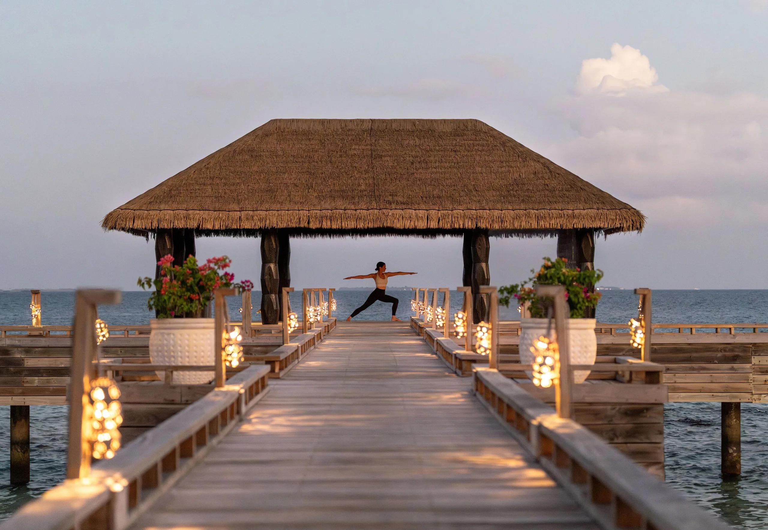 A guest practising yoga at dusk on the lantern-lit overwater pavilion of JOALI's JOALI BEING Cure wellness centre